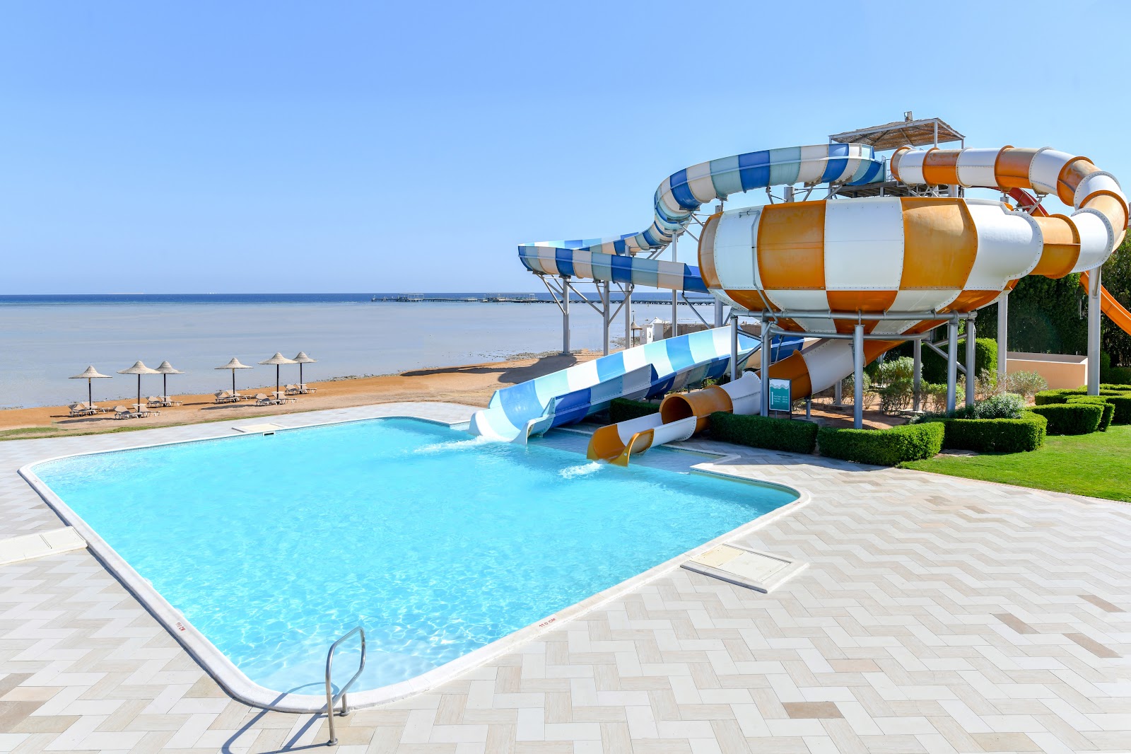 Turquoise pool with blue and orange water slides next to beach umbrellas and calm sea beyond at Jaz Aquamarine Resort.