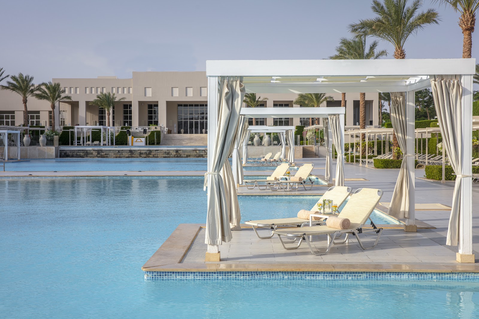 Cabanas with cream curtains frame lounge chairs beside a large pool at JAZ Aquaviva, with the beige resort building and palm trees beyond.