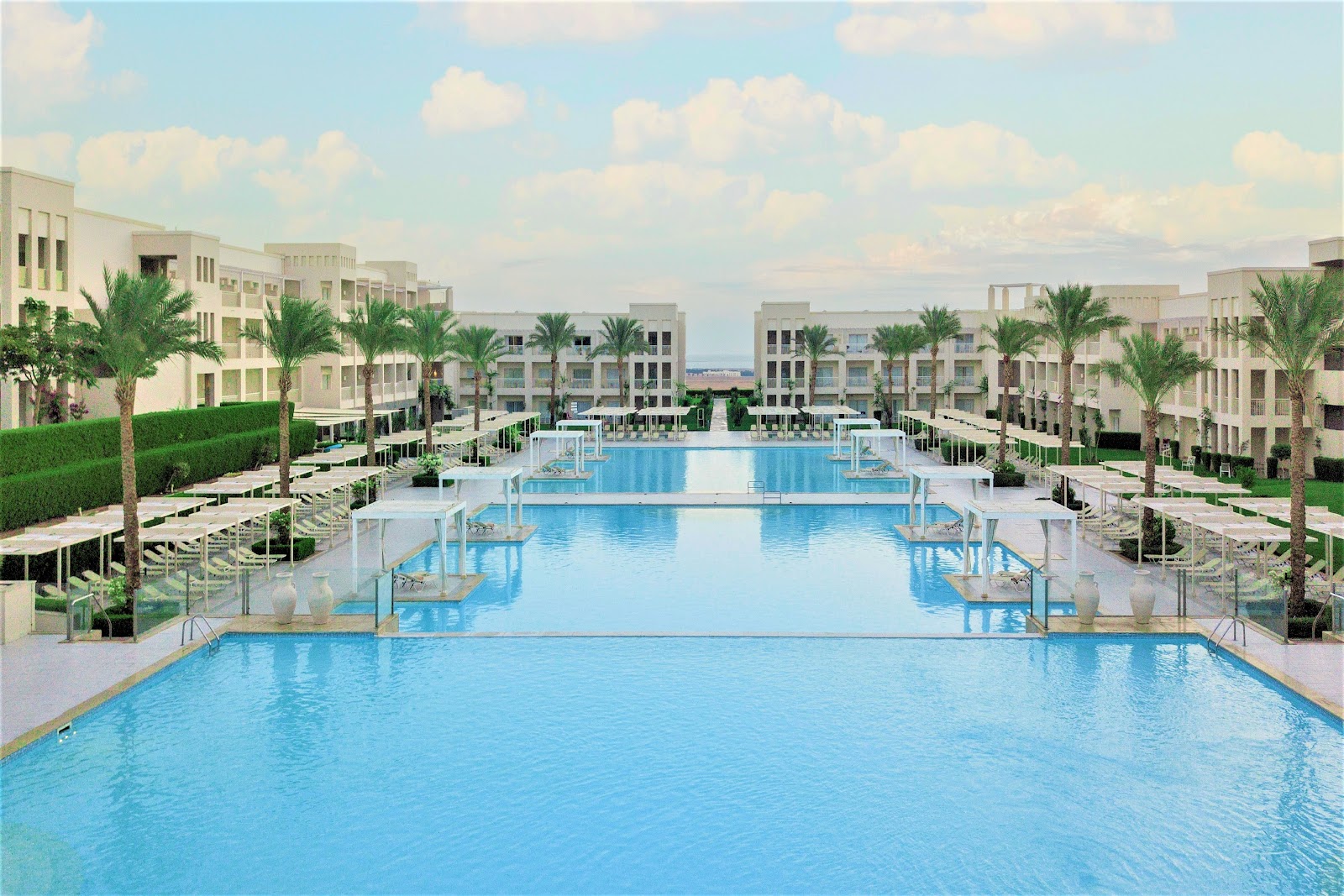 Multiple connected swimming pools with loungers and umbrellas, flanked by cream-colored resort buildings and palm trees under a partly cloudy sky.