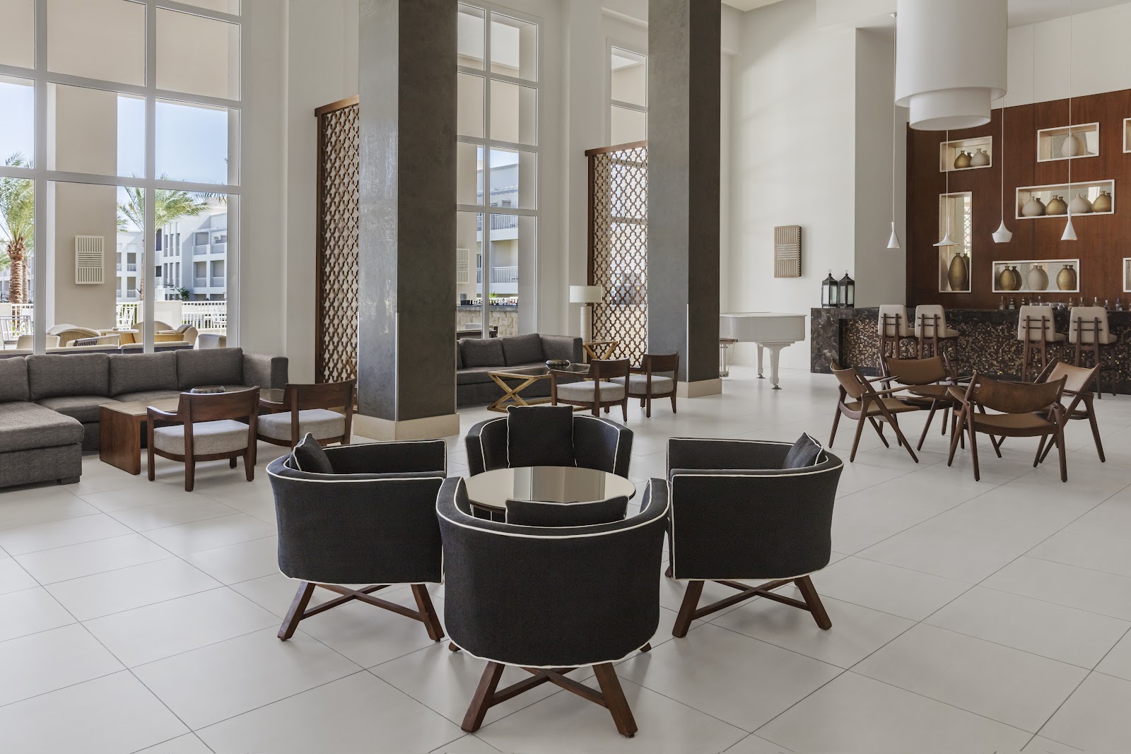 Modern lobby with white tile floor, black curved chairs arranged in center, gray sofas, dark columns, and large windows overlooking palm trees and resort buildings.
