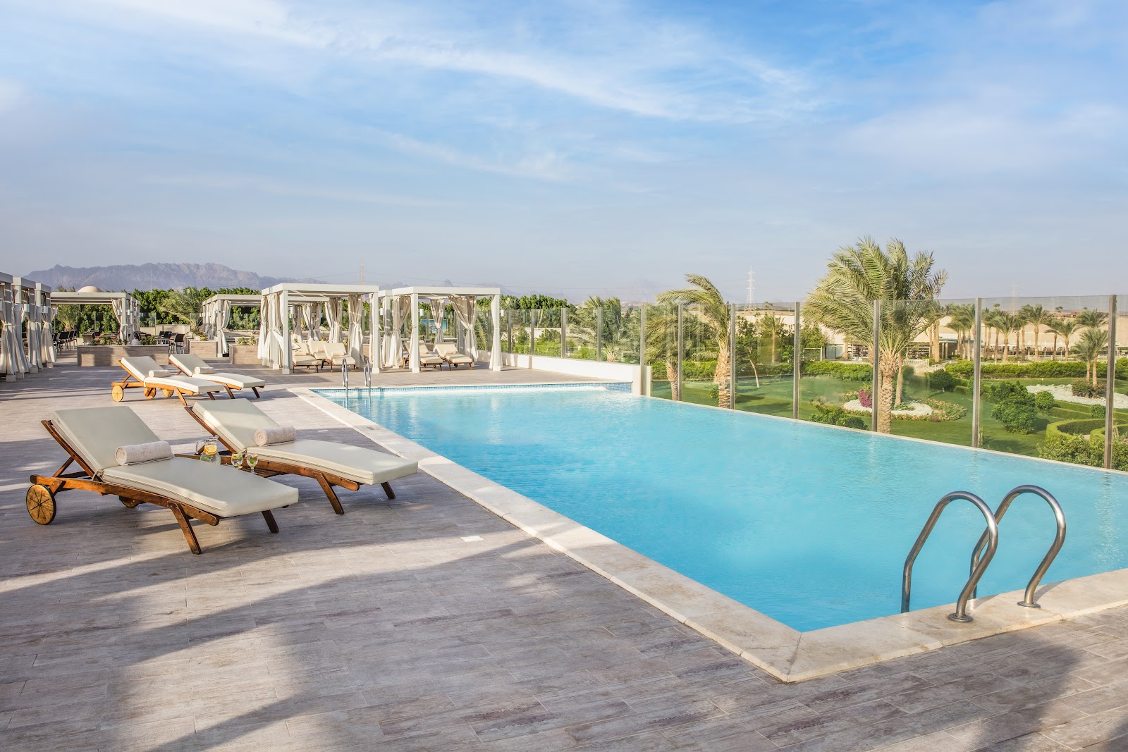 Rectangular blue pool with lounge chairs and cabanas on a paved deck overlooking palm-lined grounds and desert landscape at JAZ Aquaviva.
