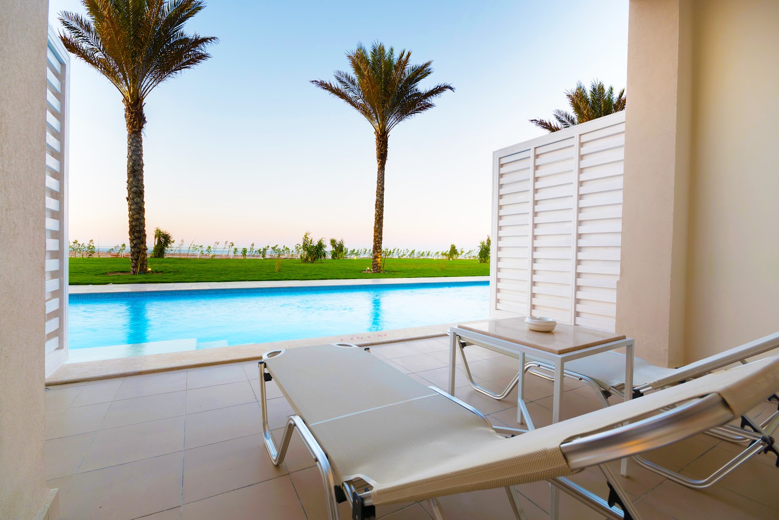 Lounge chairs on a patio overlooking a rectangular pool, palm trees, and manicured lawns under clear sky at JAZ Aquaviva.