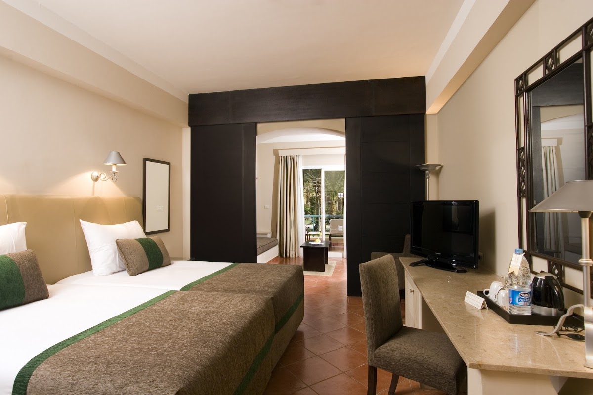 Resort room with two beds, dark wood archway, desk with chair, TV, and glass doors opening to patio with ocean view.