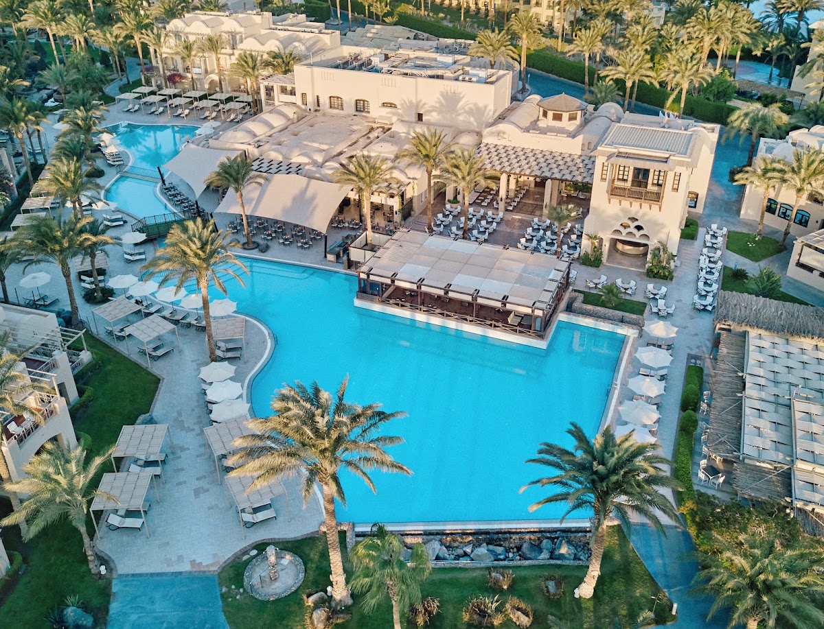 Aerial view of JAZ Makadina's large turquoise pool surrounded by cream-colored buildings, white loungers, palm trees, and manicured lawns.