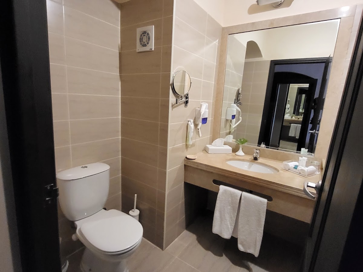 Modern bathroom with white toilet, tan tile walls, wooden vanity with white sink, large mirror, and folded towels on rack.