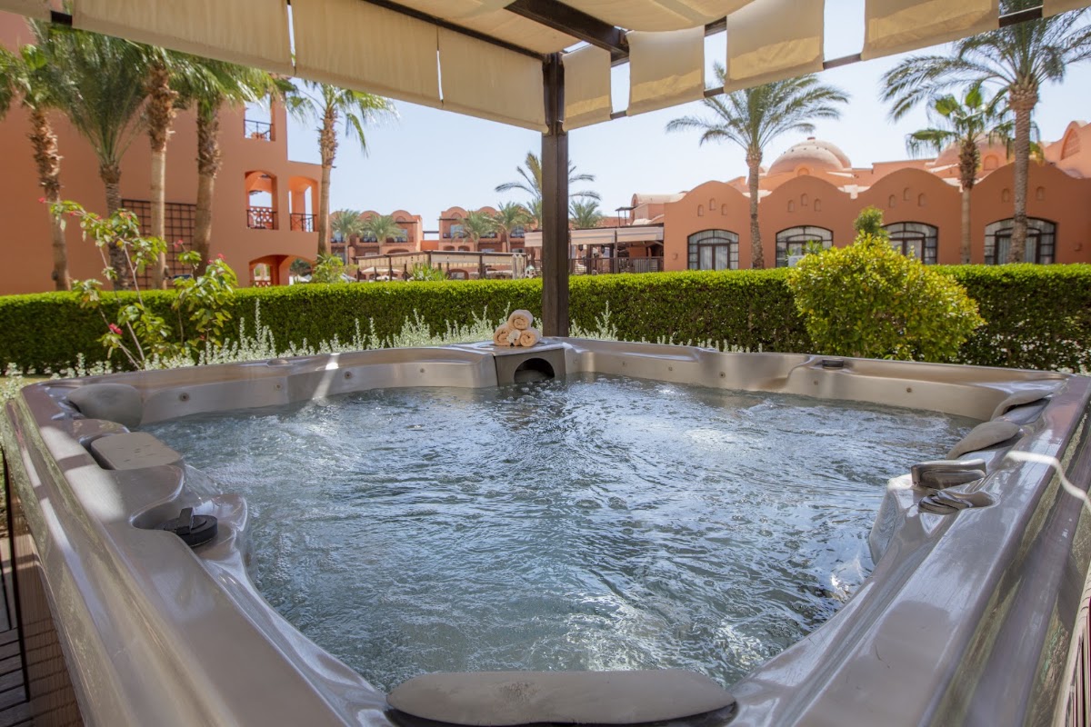 Hot tub with built-in seats under a beige canopy, framed by palm trees and terracotta resort buildings in a desert setting.