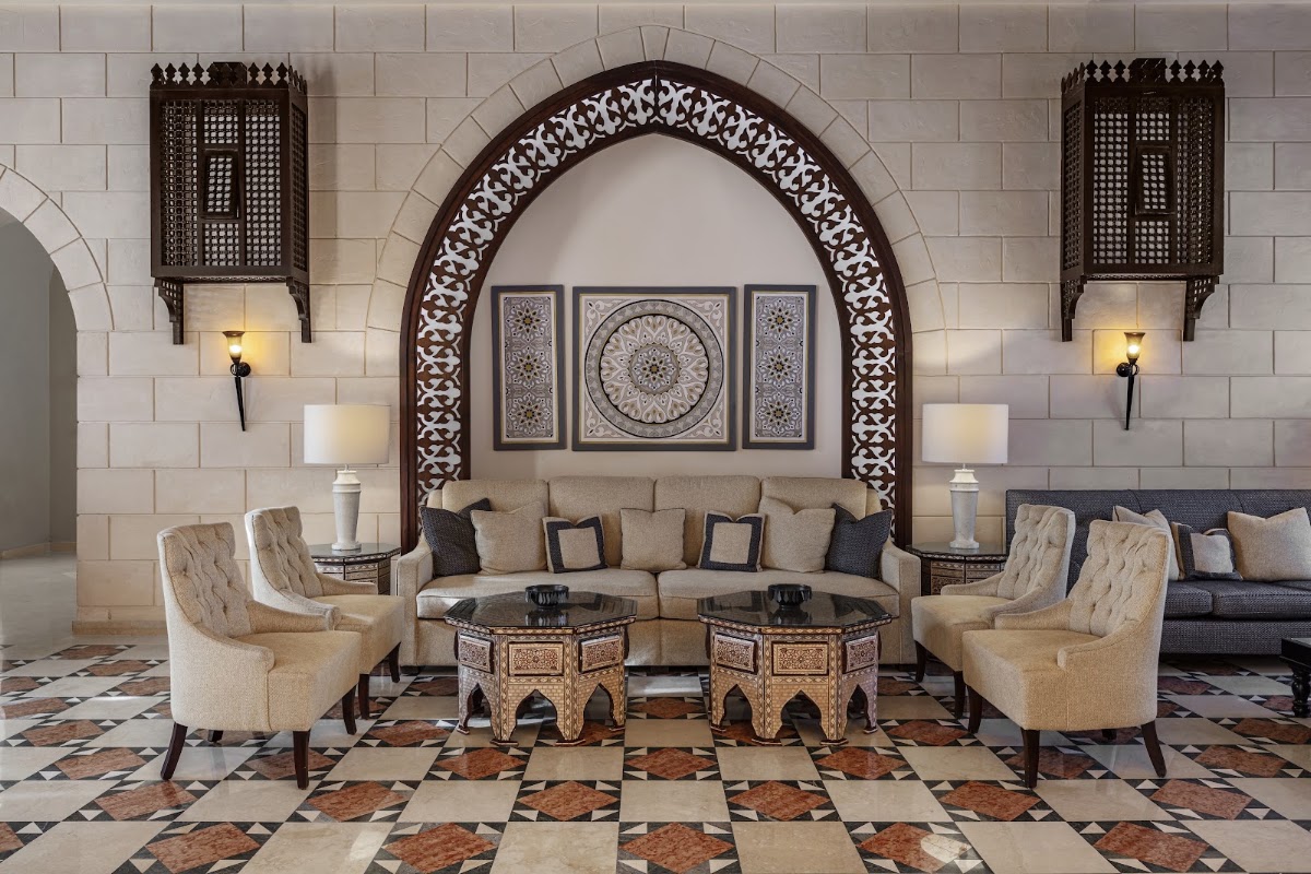 Moroccan-style lounge with cream sofa, beige armchairs, carved wooden arch, ornamental wall panels, and geometric tile floor.