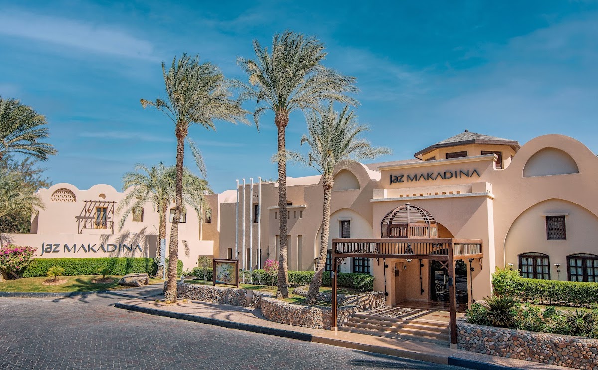 Terracotta-colored buildings with arched windows at JAZ Makadina, framed by tall palm trees under clear blue sky.