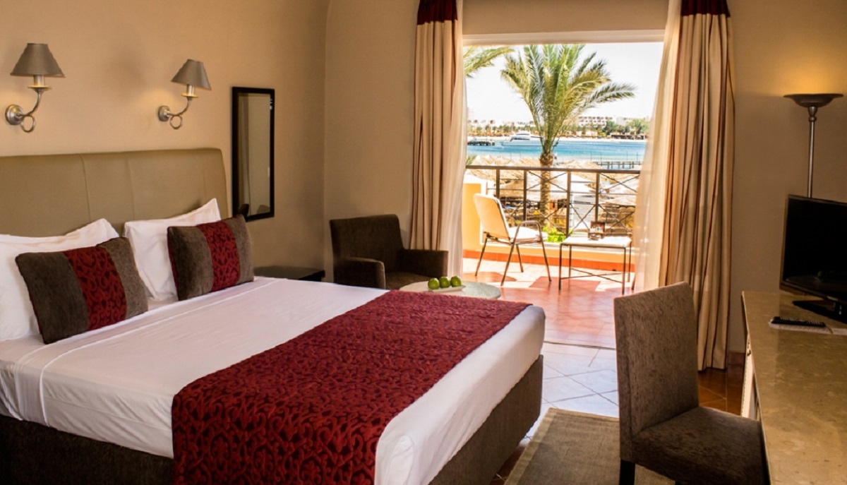 Hotel room with a queen bed, balcony overlooking a palm tree and blue waterway, and neutral beige walls with burgundy accents.