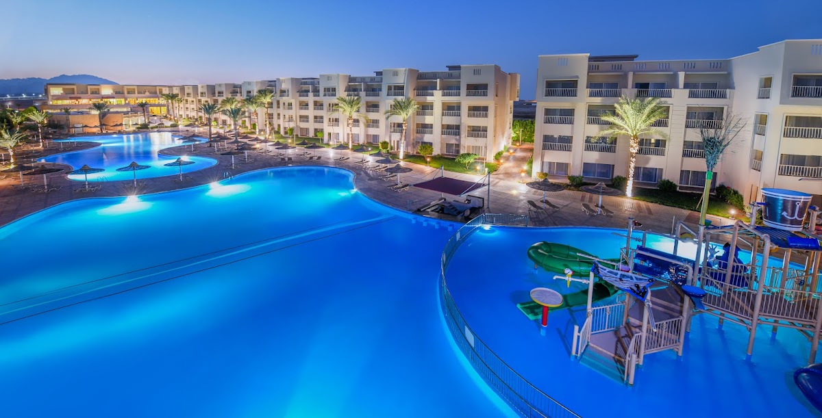 Illuminated pools with water slides and playground equipment at dusk, bordered by multi-story resort buildings and palm trees.