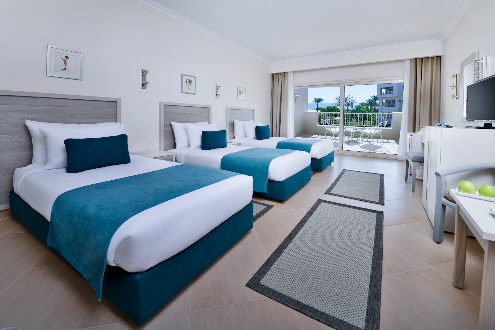Resort room with three beds in teal and white, balcony overlooking palm trees and ocean, TV mounted on wall.