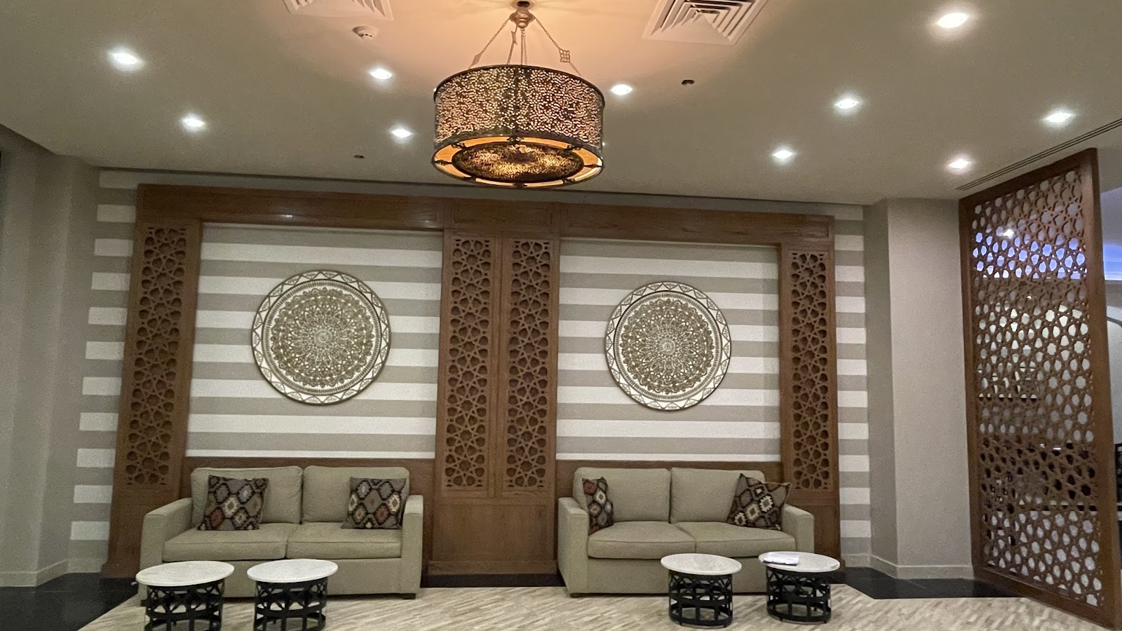 Spa lounge at JAZ Soma Beach with beige sofas, ornate wooden wall panels, decorative brass pendant light, and patterned marble side tables.