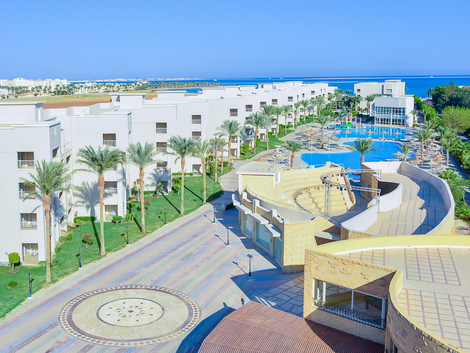 Rooftop lounge area with white cushioned seating overlooking a wooden deck, decorative circular pattern, palm trees, and resort buildings under clear blue sky.