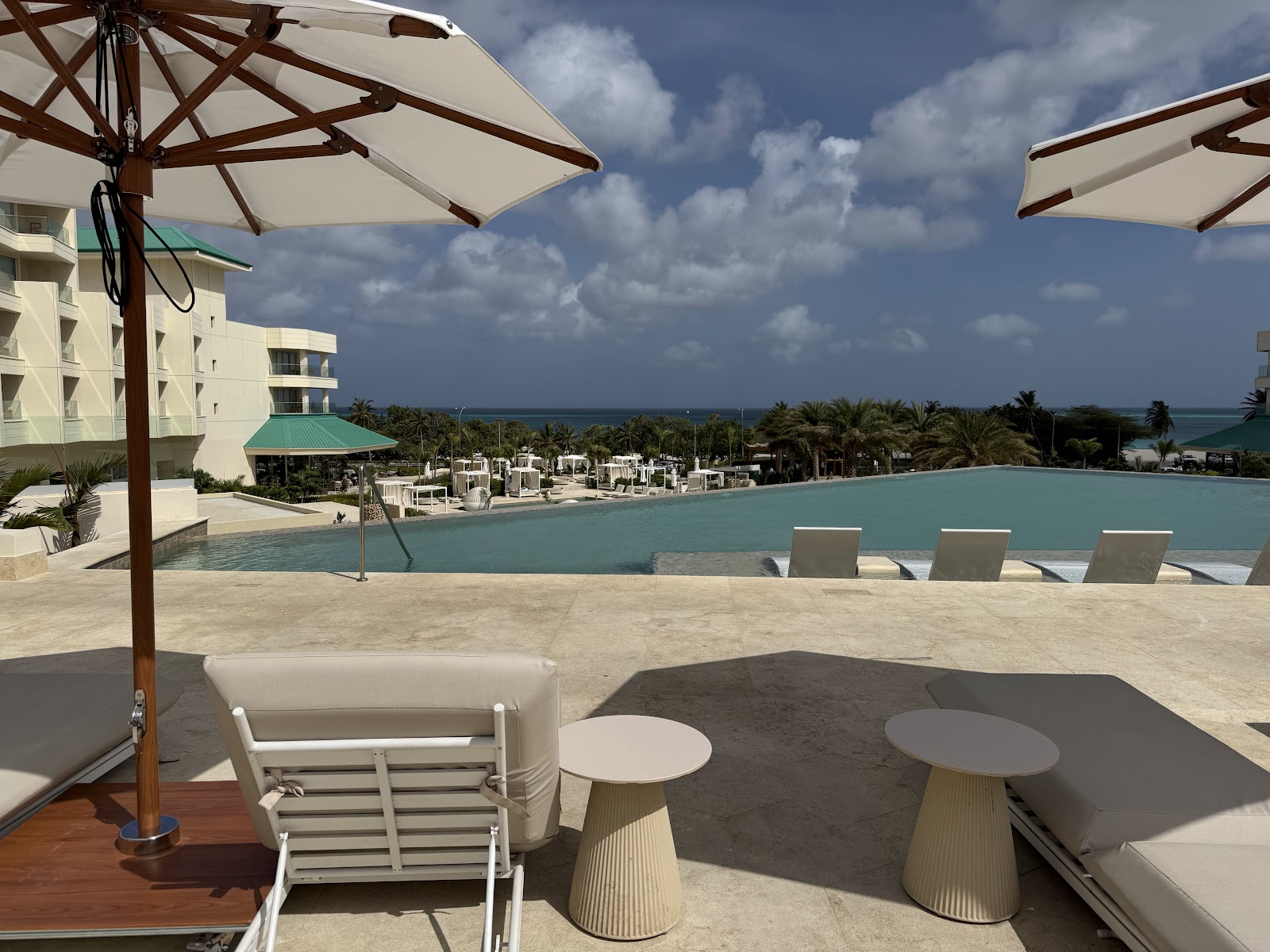 Poolside lounge area at JOIA Aruba by Iberostar with cream umbrellas, white loungers, and ocean view beyond palm trees.