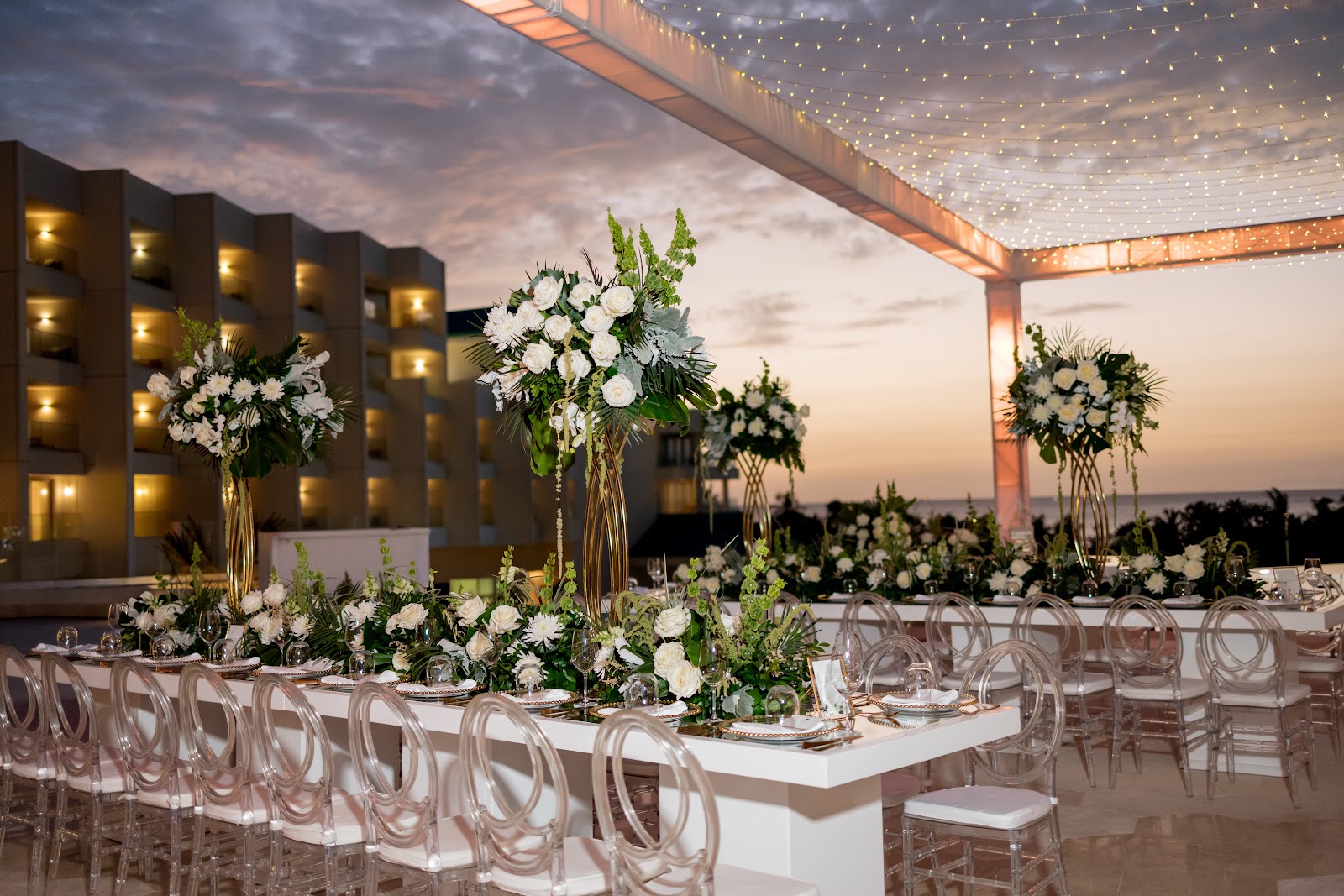 Outdoor event space at dusk with white floral centerpieces, transparent chairs, and string lights overhead, resort building lit in background.