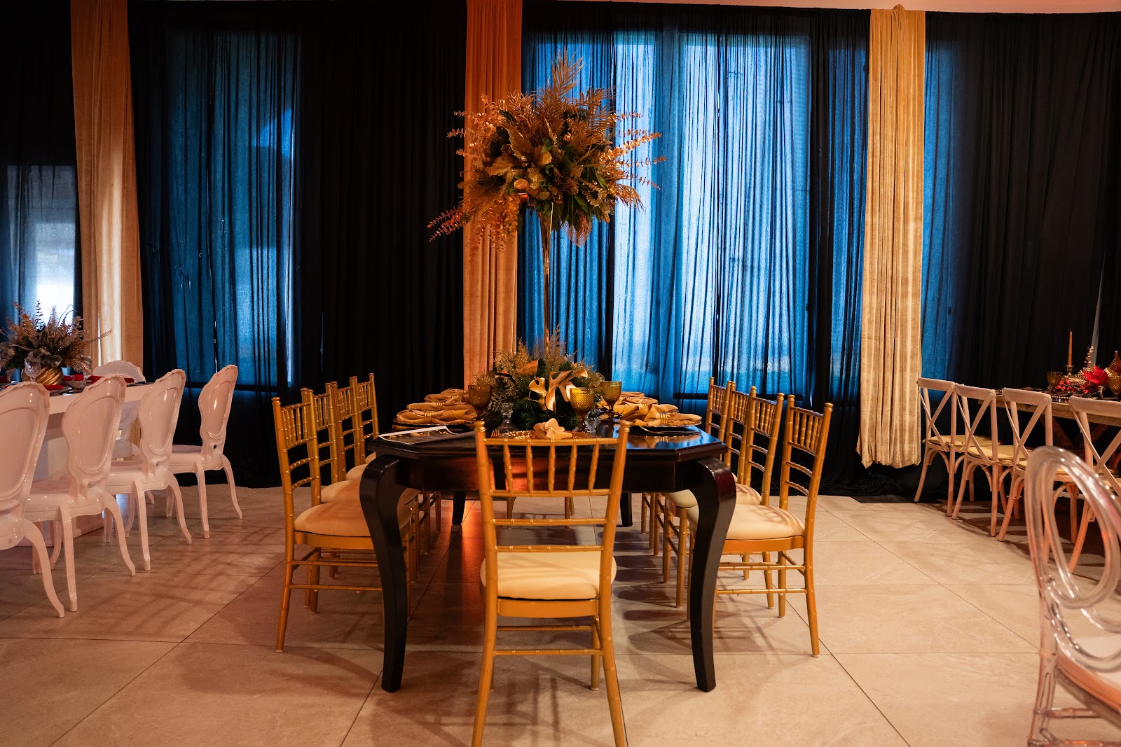 Banquet hall with round black table, wooden chairs, tall floral centerpiece, and blue curtains lining the windows.