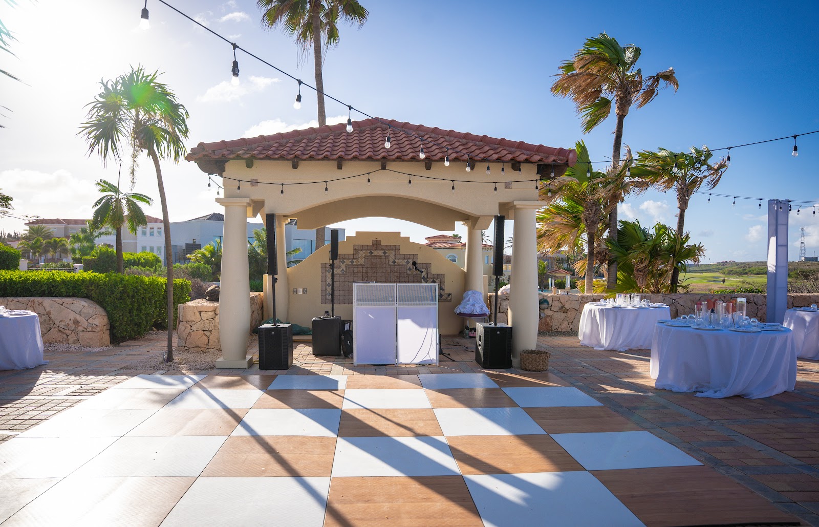 Outdoor event space at JOIA Aruba by Iberostar with arched pavilion, checkered tile floor, white-clothed tables, palm trees, and ocean views beyond.