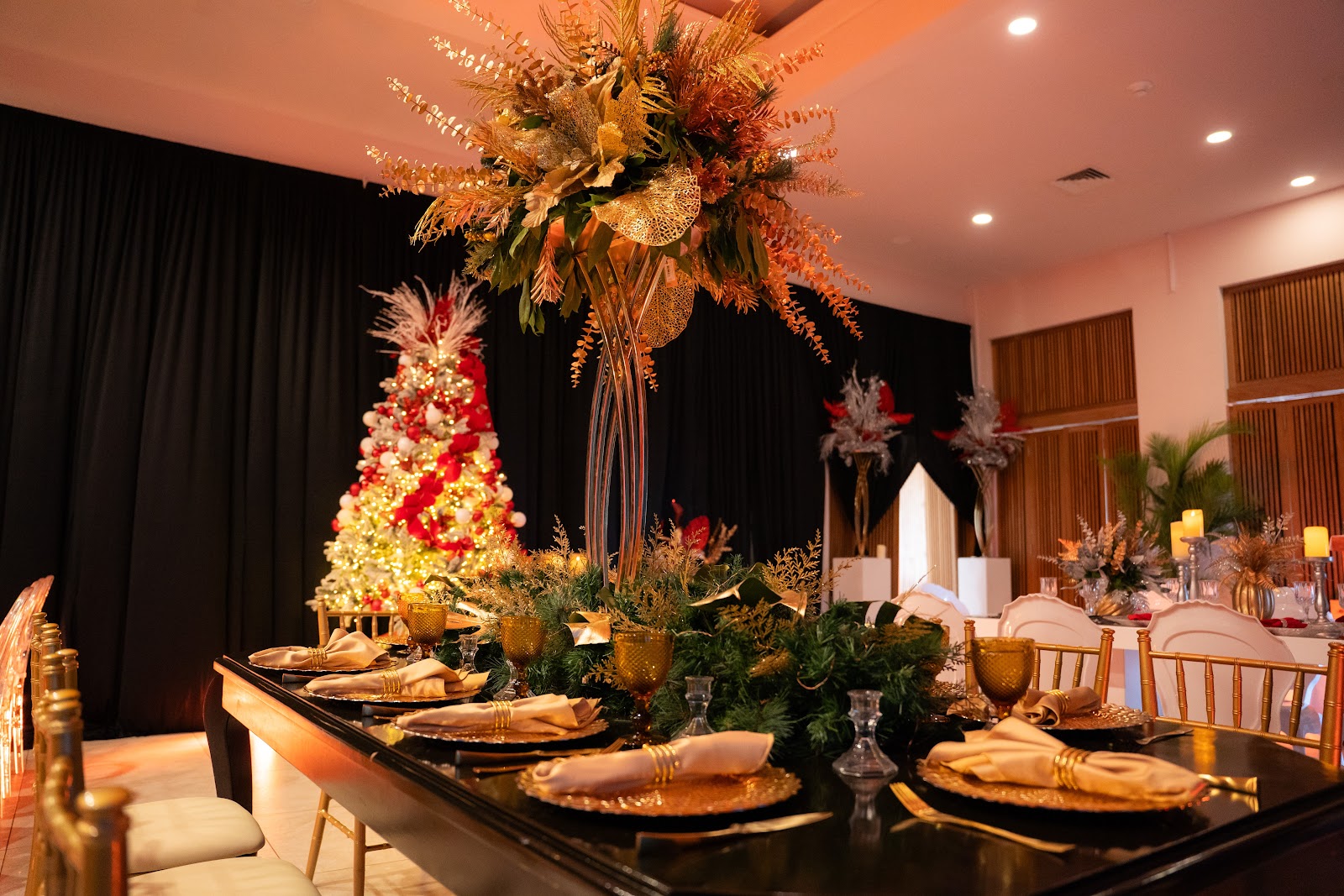 Elegant ballroom with a black banquet table set with gold place settings, tall floral centerpiece with dried grasses overhead, and a red and gold Christmas tree against black curtains.