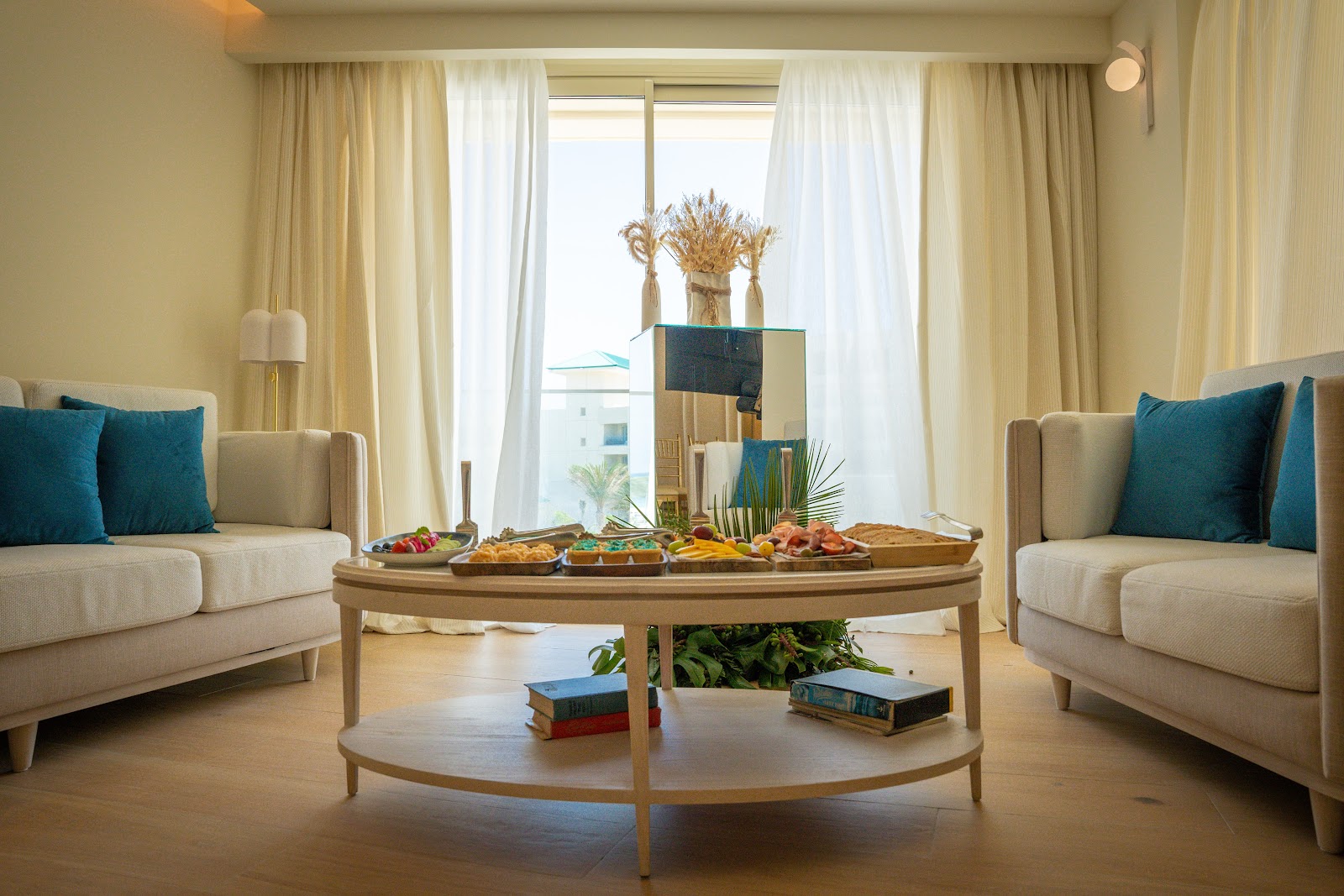 Resort suite with cream sofas flanking a wooden oval table displaying appetizers and flowers, floor-to-ceiling windows overlooking ocean views.