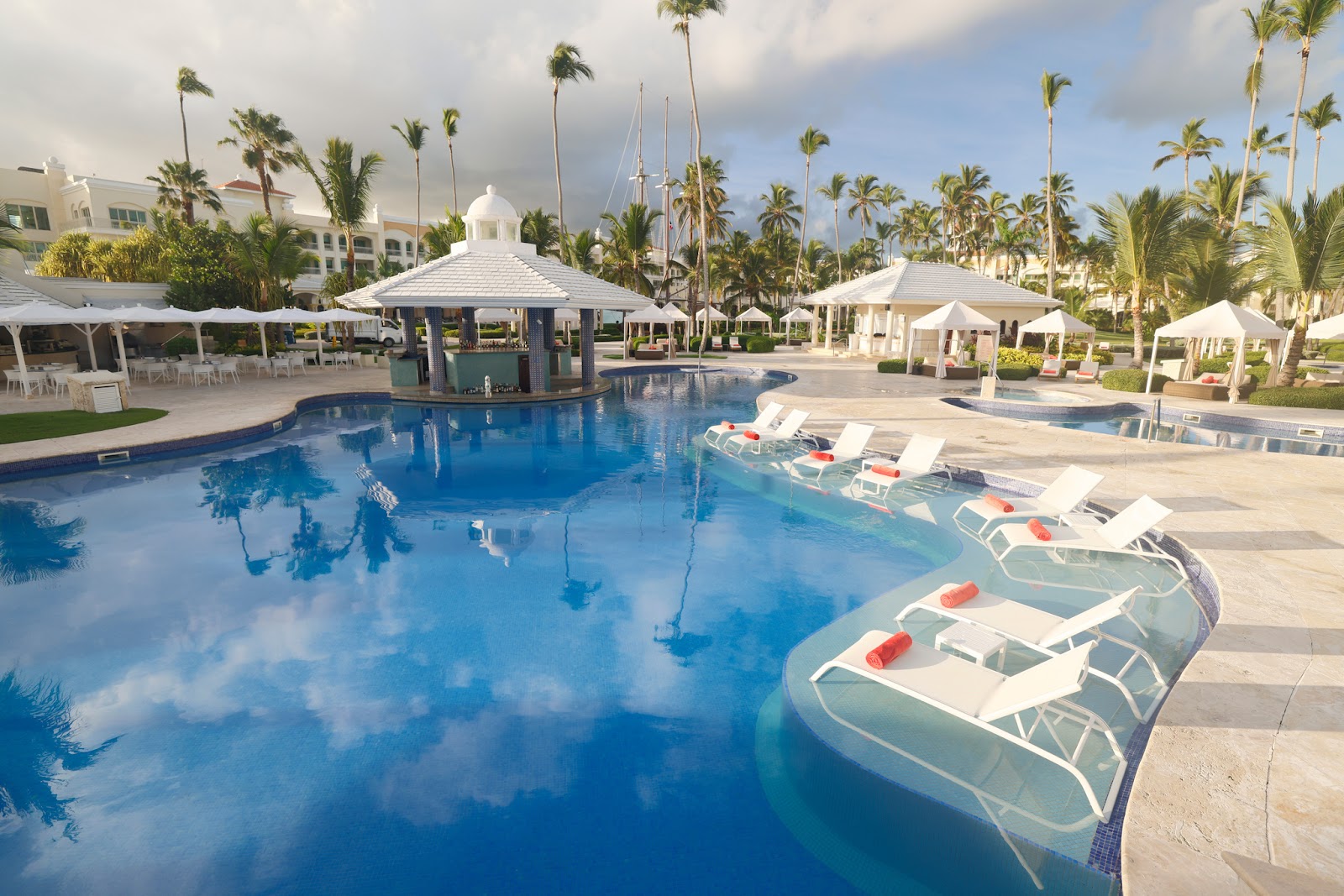 Resort pool with white umbrellas, loungers with red towels, a central gazebo bar, palm trees, and white buildings under partly cloudy sky.