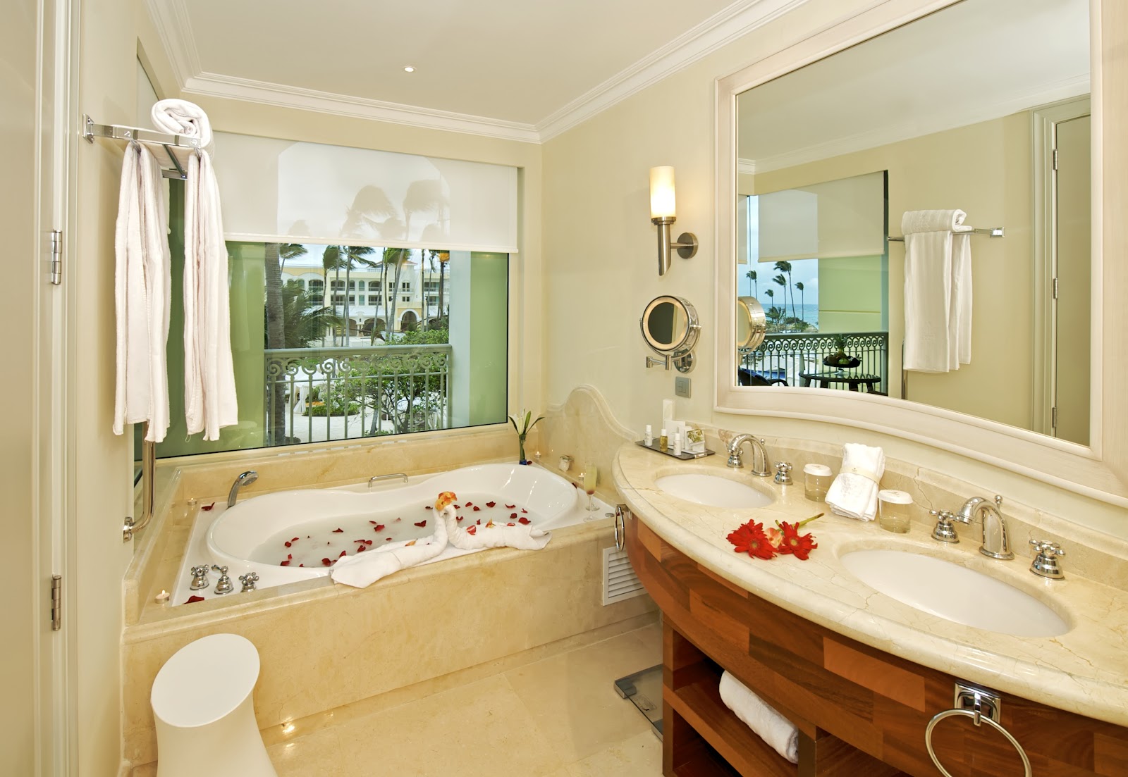 Bathroom with cream walls and marble surfaces, soaking tub with red flower petals overlooking a balcony with palm trees and resort grounds.
