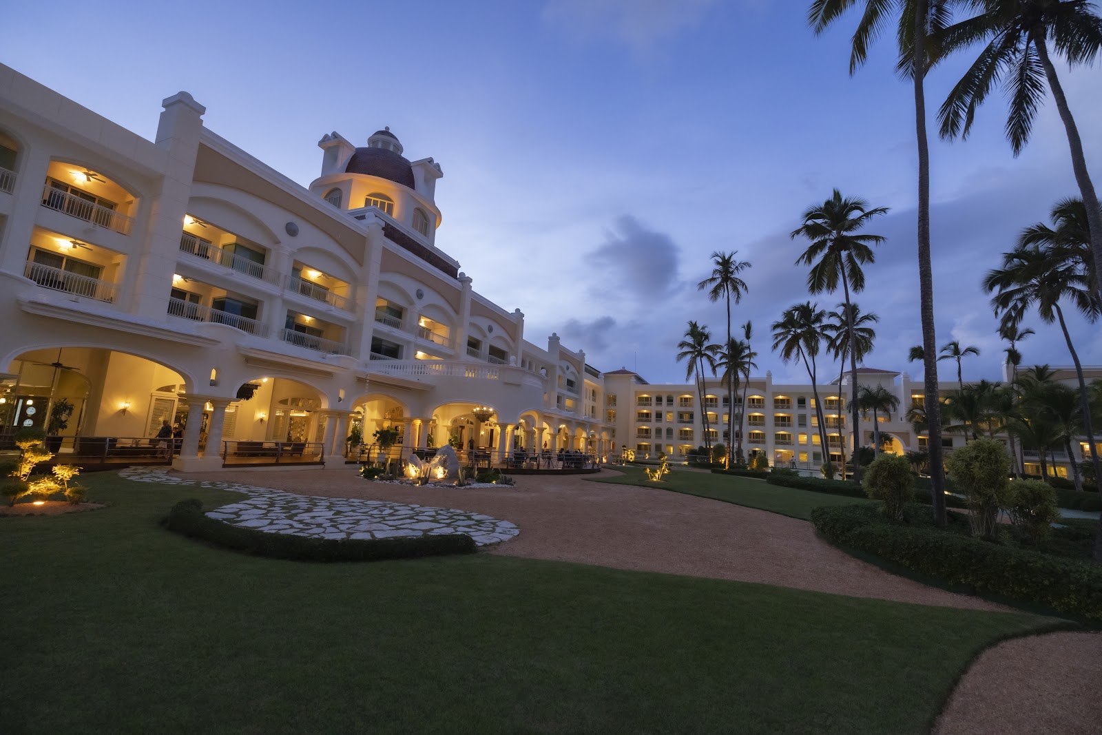 Spacious resort suite with cream walls, dark wood furniture, flat-screen TV, and glass doors opening to a balcony overlooking palm trees.
