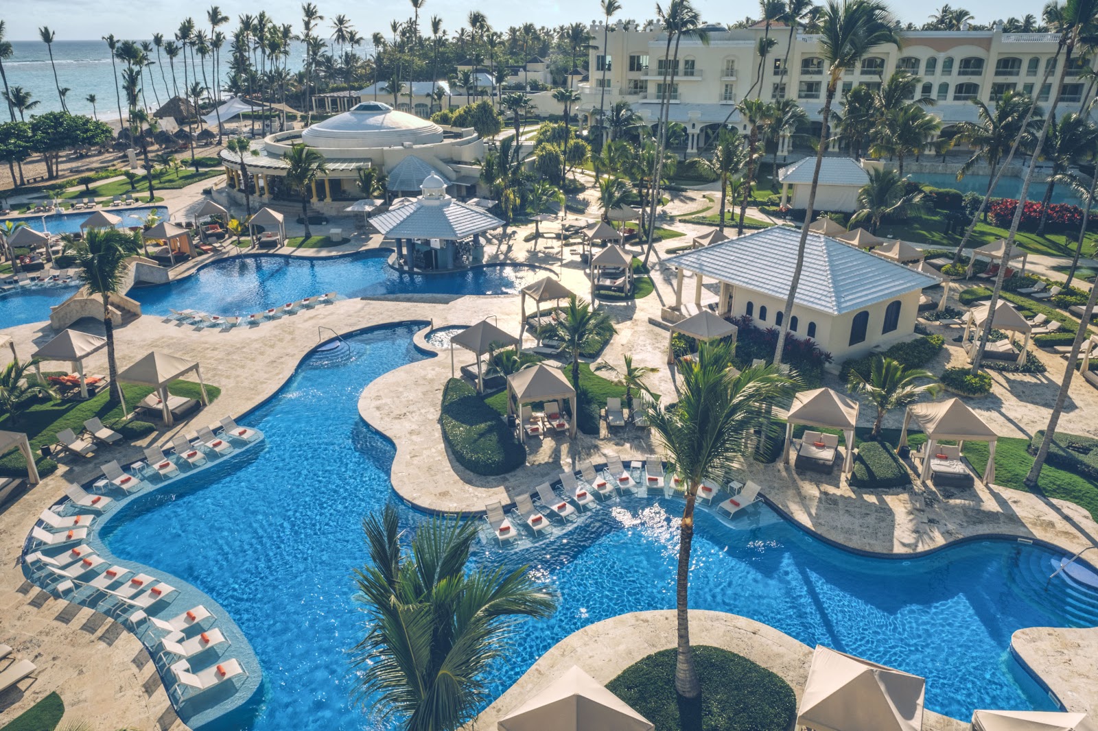 Aerial view of JOIA Bávaro by Iberostar with blue pools, white buildings, palm trees, and a turquoise lagoon opening to a sandy beach and ocean.
