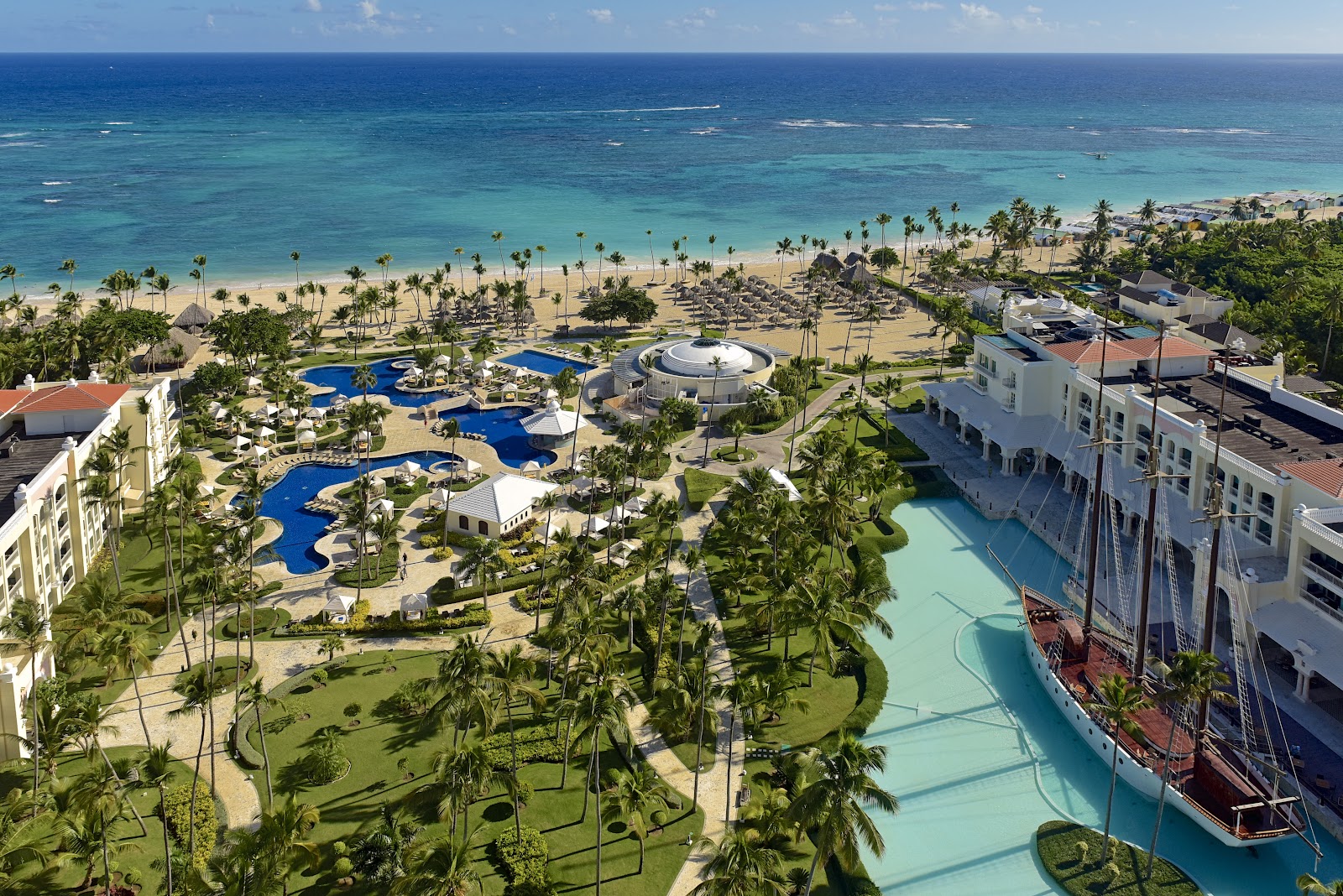 Aerial view of JOIA Bávaro by Iberostar beachfront resort with white buildings, pools, and rows of lounge chairs on sand facing turquoise ocean waves.