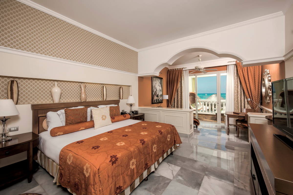 Beachfront resort bedroom with ocean view through glass doors, queen bed with orange patterned bedding, and neutral wall accents.