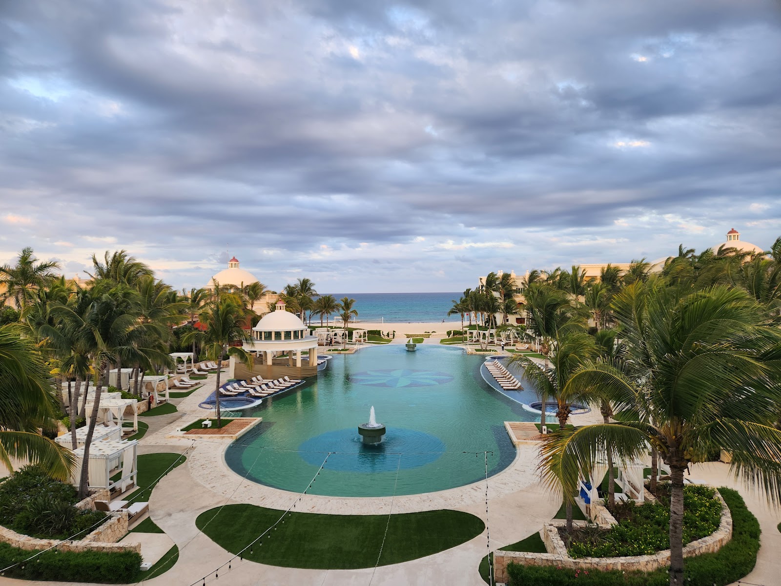 Lounge chairs on a lawn face a large turquoise lagoon with a fountain, bordered by yellow colonial-style buildings and palm trees under blue sky.