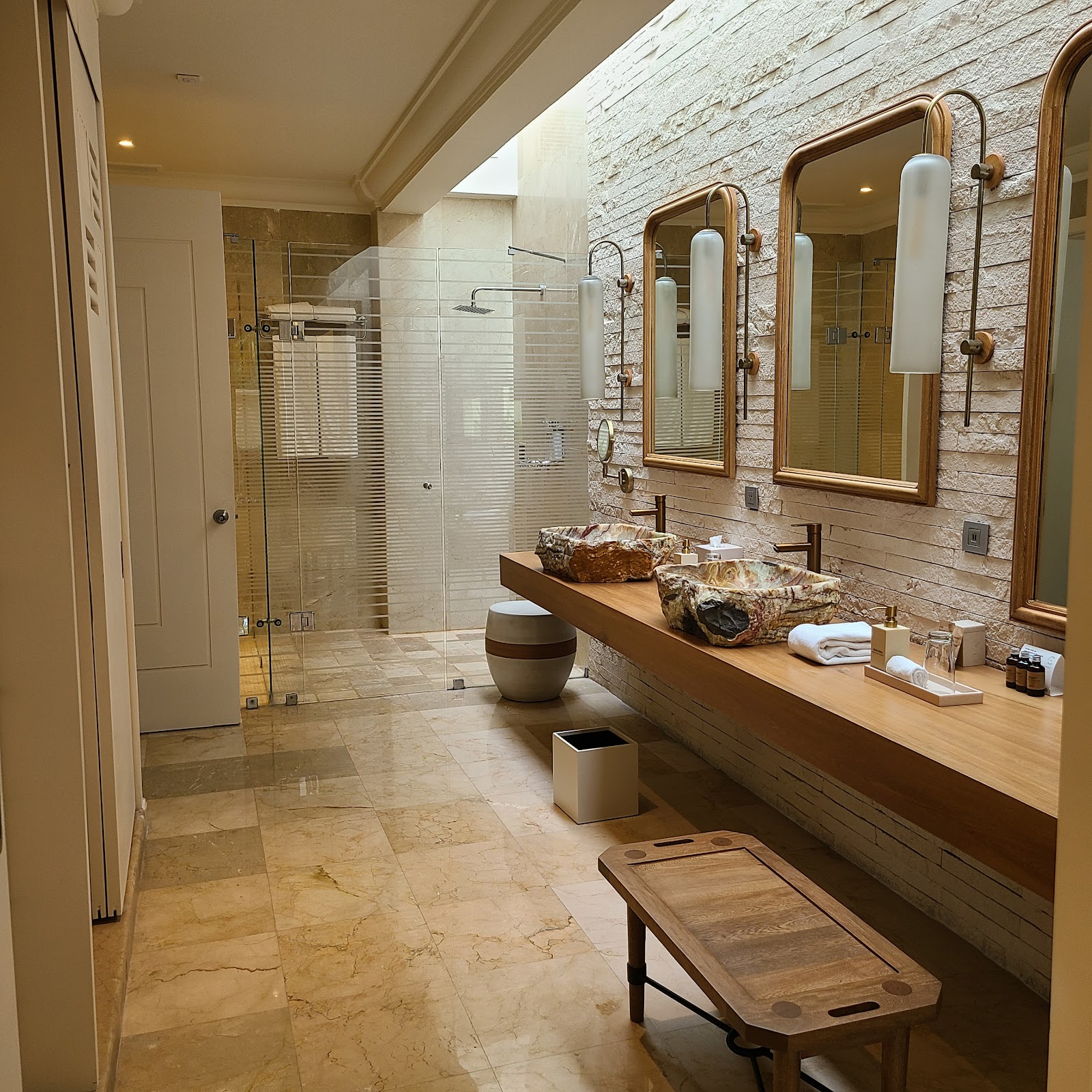 Spacious bathroom with dual vanity, stone accent wall, framed mirrors, walk-in shower, and marble flooring at JOIA Paraíso by Iberostar.