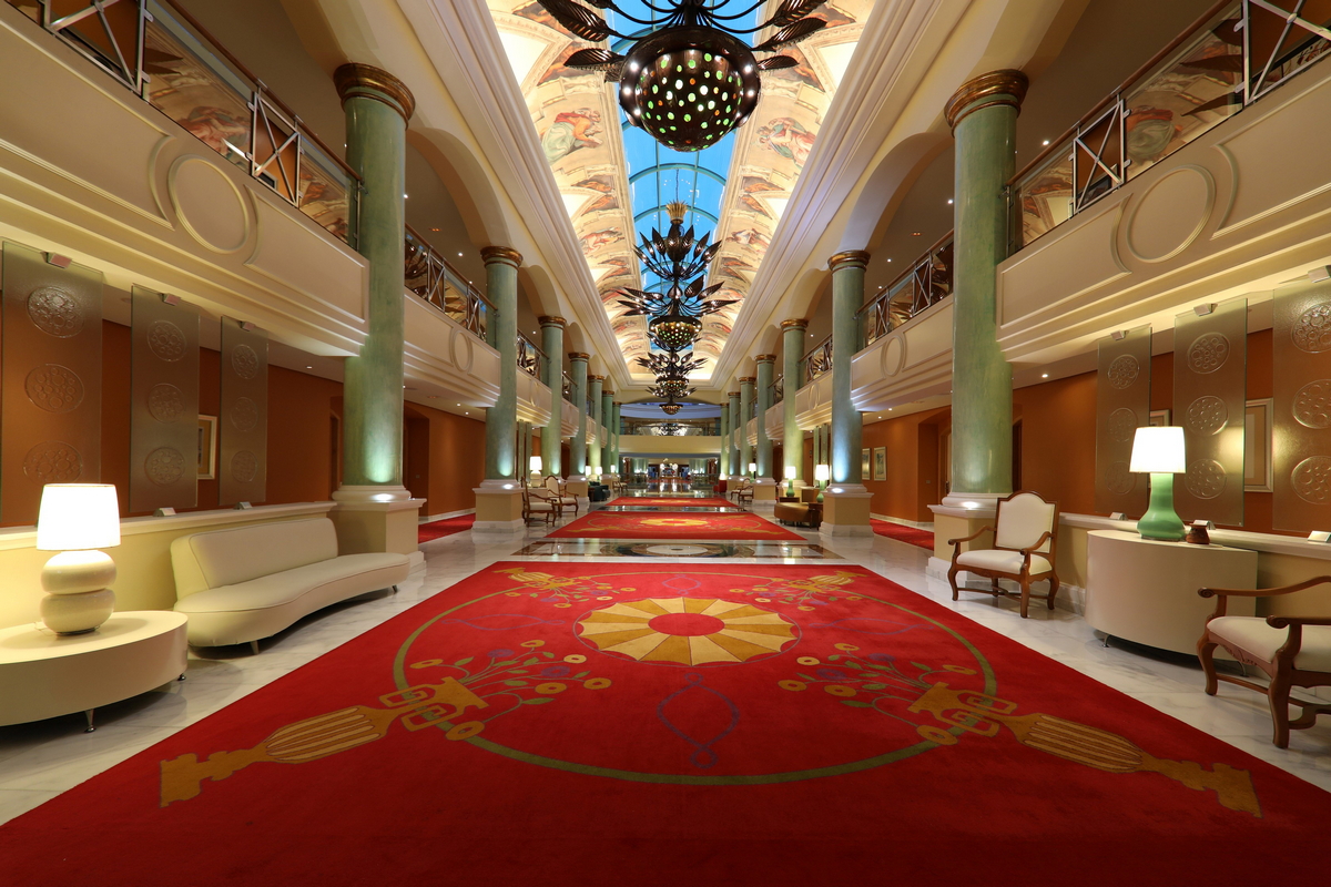 Grand lobby hallway with red patterned carpet, green columns, cream-colored walls, and ornate ceiling fixtures at JOIA Paraíso by Iberostar.