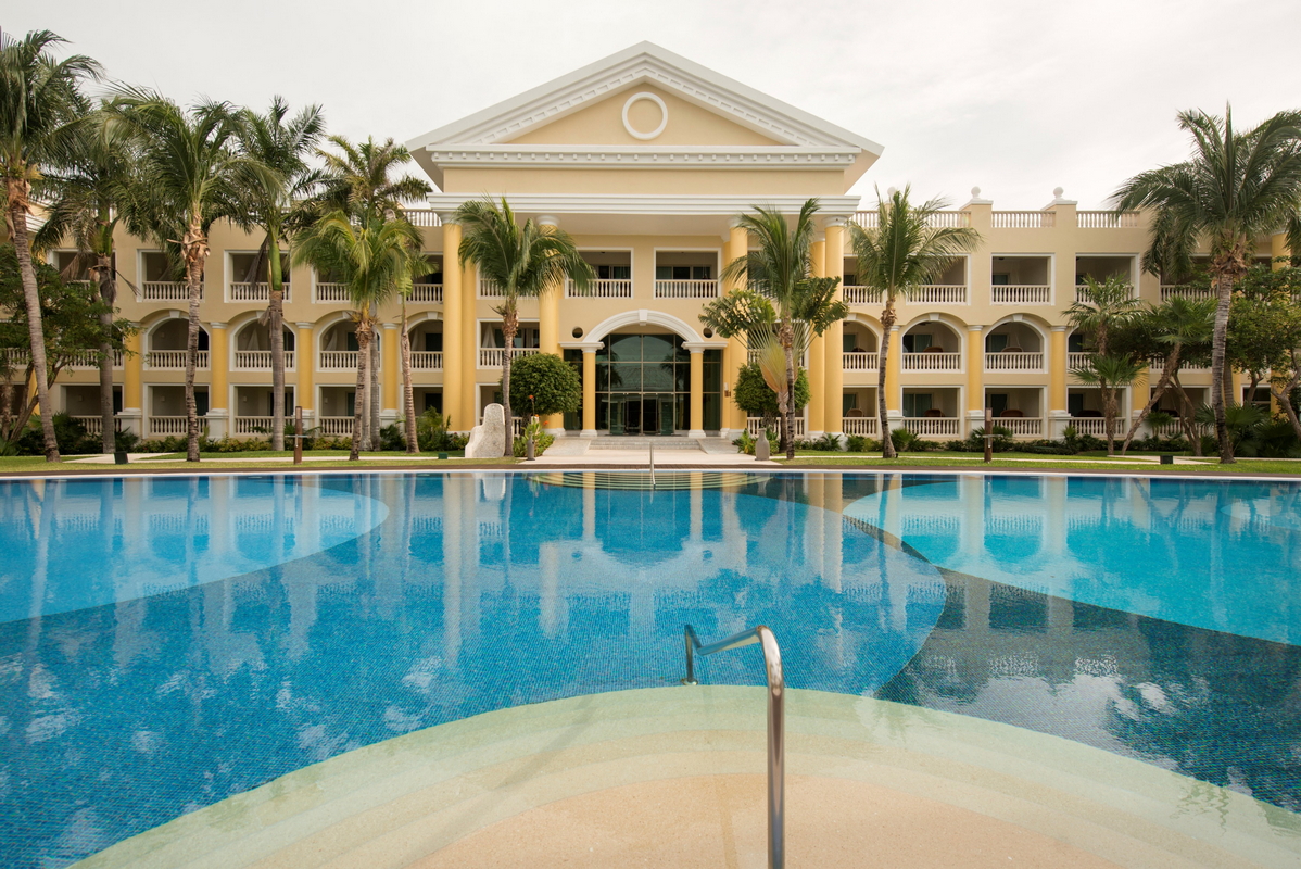 Resort pool with turquoise water and fountains facing pale yellow Mediterranean-style main building with domed roof and the ocean beyond.