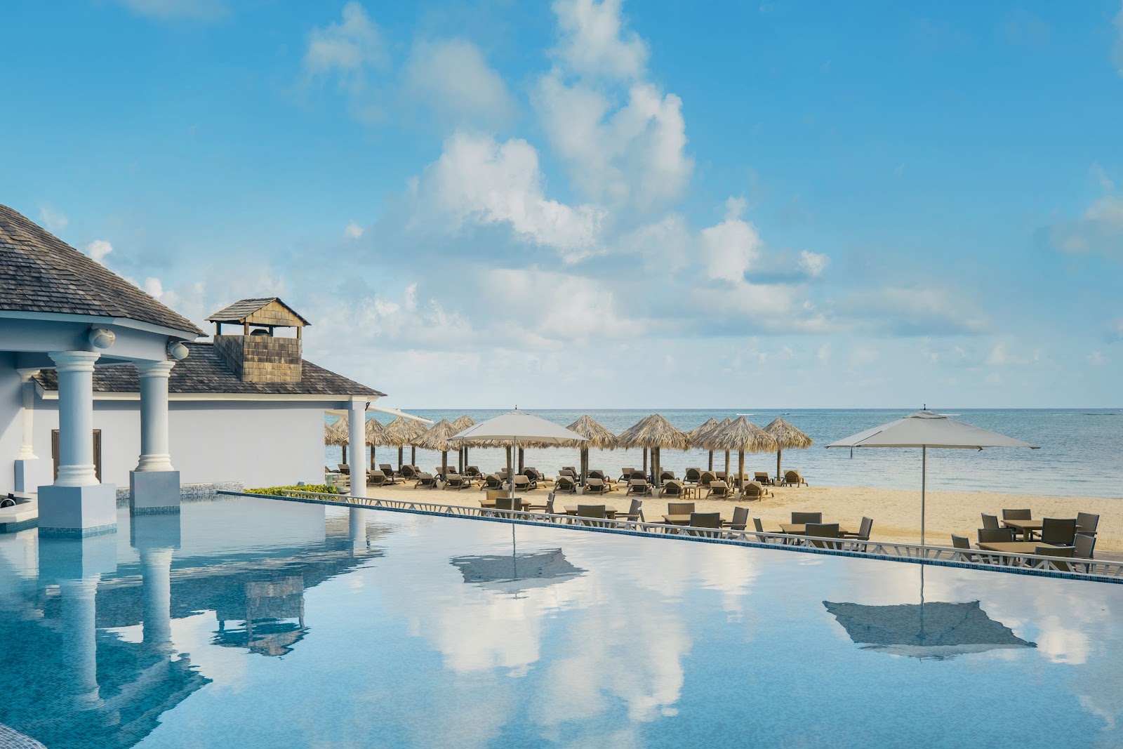 Infinity pool with white columns overlooking the beach, lounge chairs, thatched umbrellas, and calm turquoise ocean under blue sky.