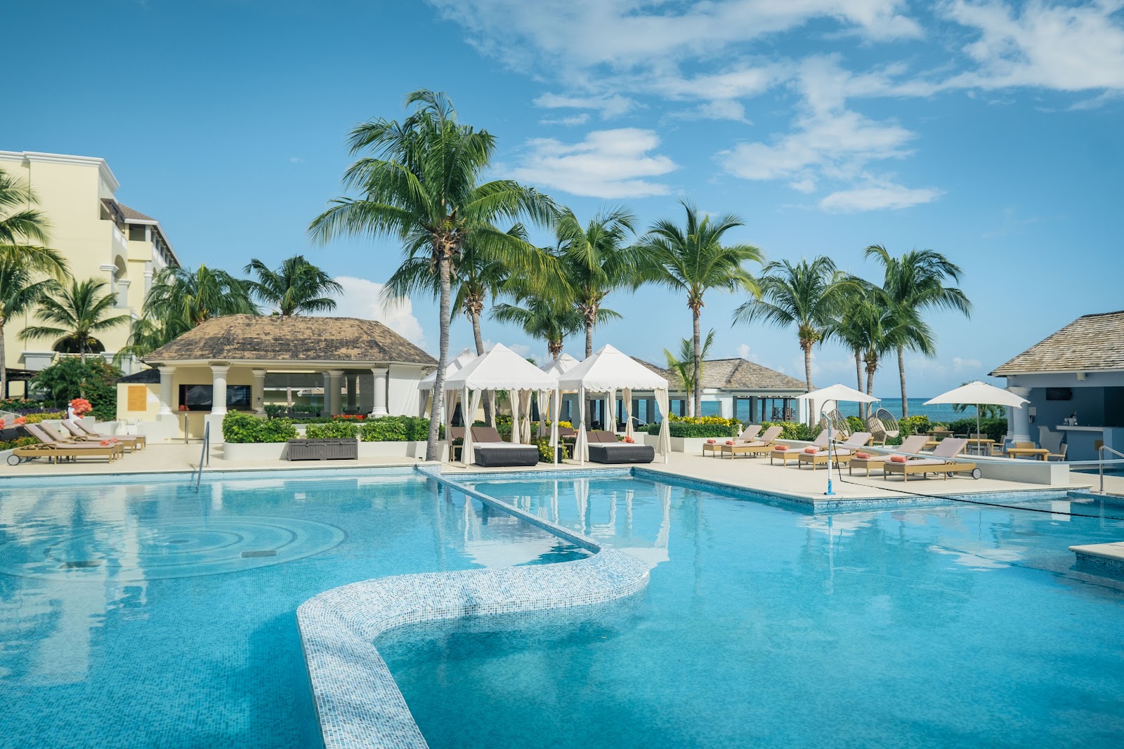 Large curved pool with white cabanas and palm trees at JOIA Rose Hall by Iberostar, ocean view in background.