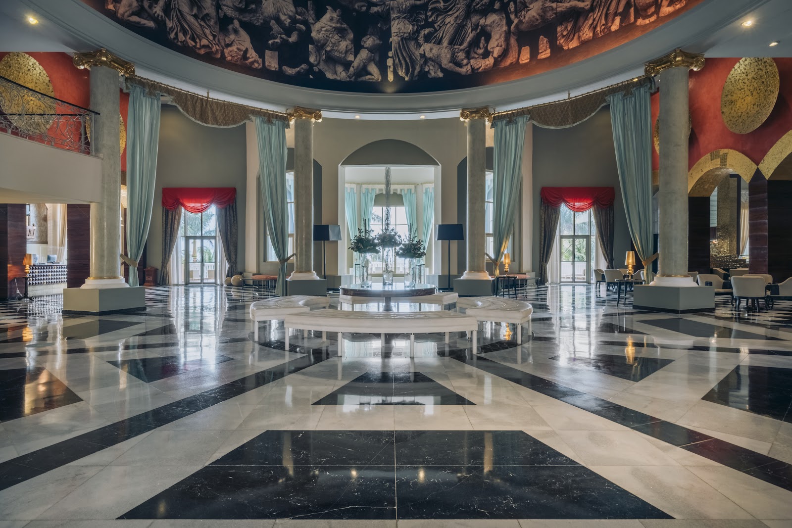 Grand lobby with polished marble floors, soaring columns, turquoise curtains, red accent walls, and ornate ceiling mural at JOIA Rose Hall by Iberostar.