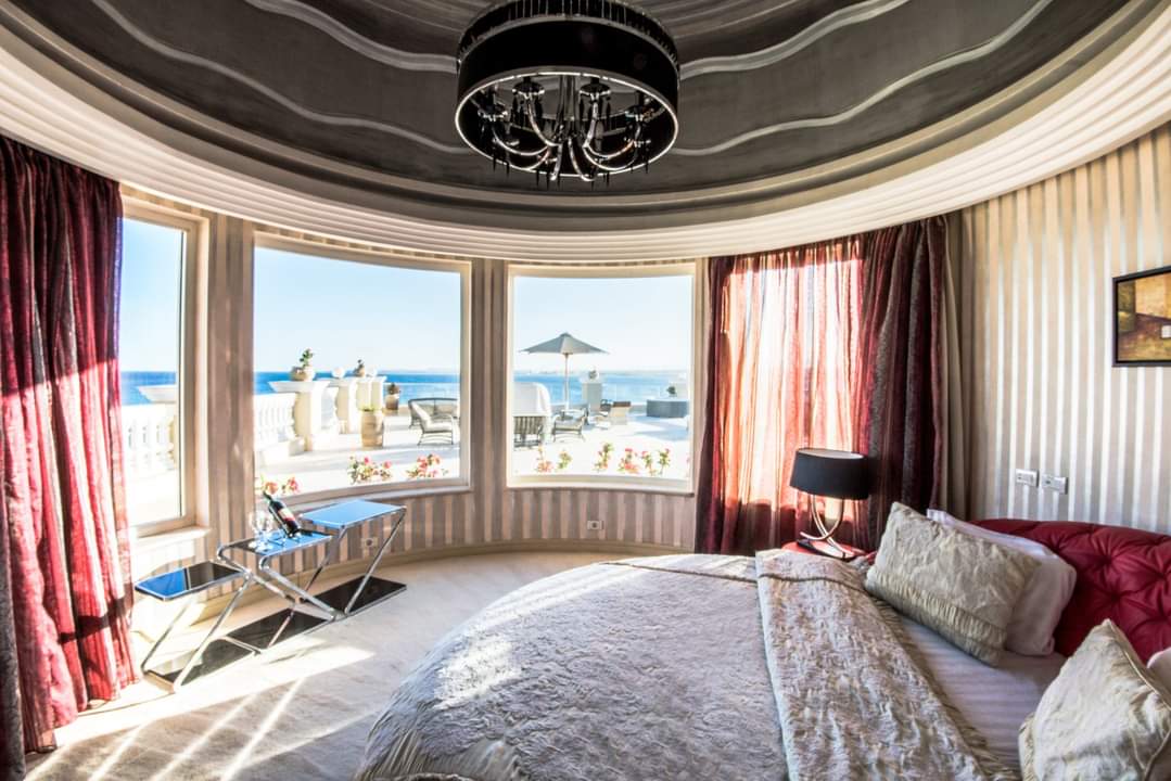 Hotel bedroom with two beds, TV, and floor-to-ceiling windows overlooking a beachside terrace and calm sea.