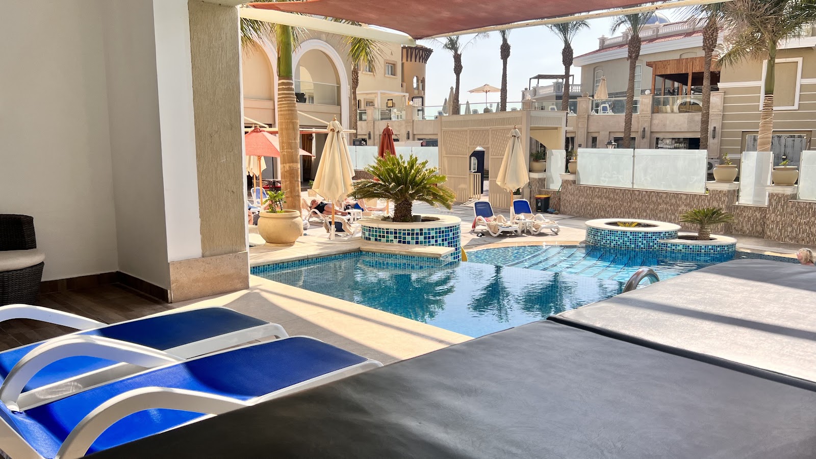 Blue loungers in foreground face a tiled pool with spa jets at KaiSol Romance Resort, surrounded by cream buildings and palm trees.