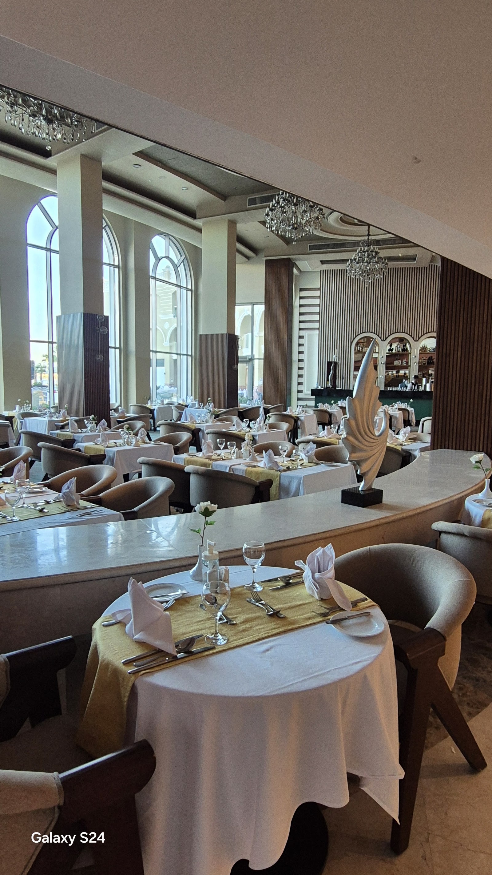 Upscale dining room with round tables set with white linens, modern curved chairs, crystal chandeliers, and floor-to-ceiling windows overlooking the coast.