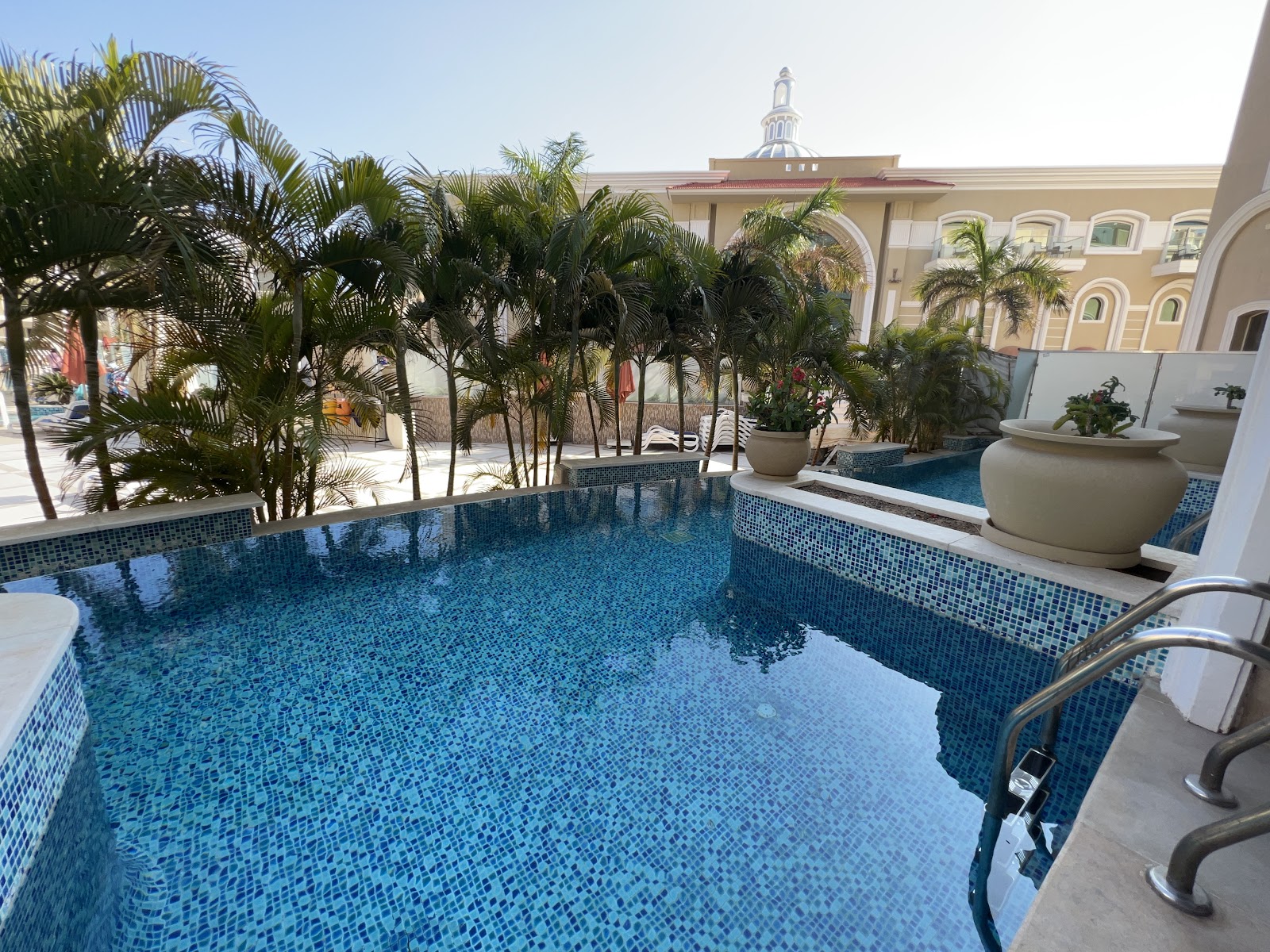 Resort pool with blue tiled walls surrounded by palm trees and tan colonial-style buildings with a cupola visible beyond.