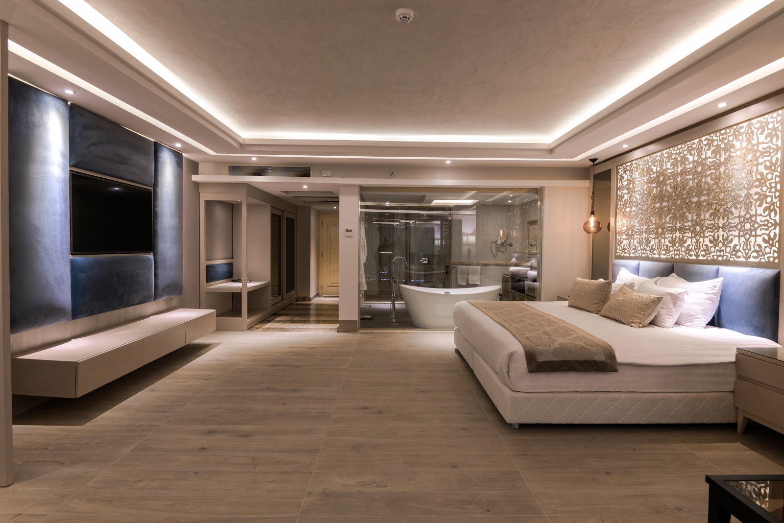 Spacious resort suite with king bed, wall-mounted TV, soaking tub visible through glass partition, and ambient ceiling lighting throughout.