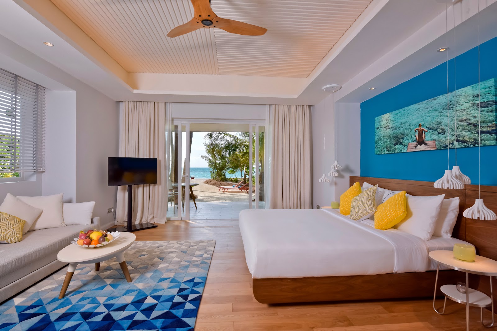 Spacious beachfront suite at Kandima Maldives with king bed, lounge area, and ocean views through glass doors to sandy beach and palm trees.