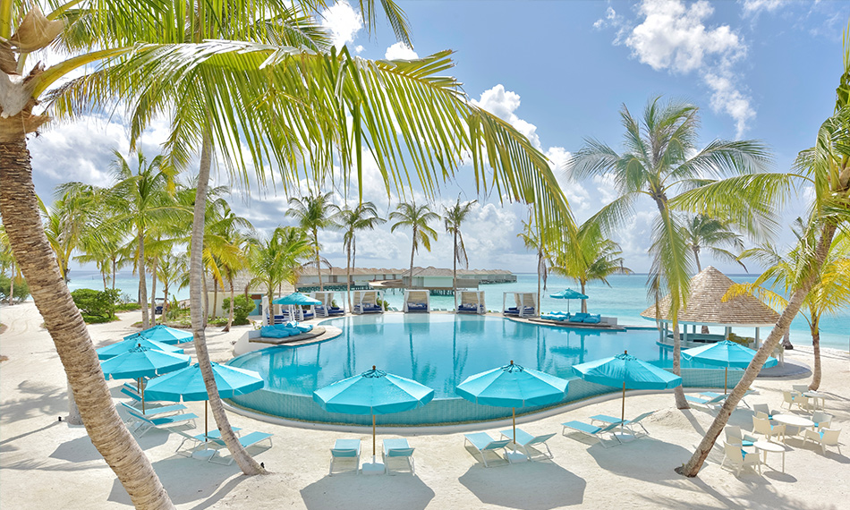 Turquoise pool with blue umbrellas and loungers surrounded by palm trees, with overwater bungalows and ocean beyond at Kandima Maldives.