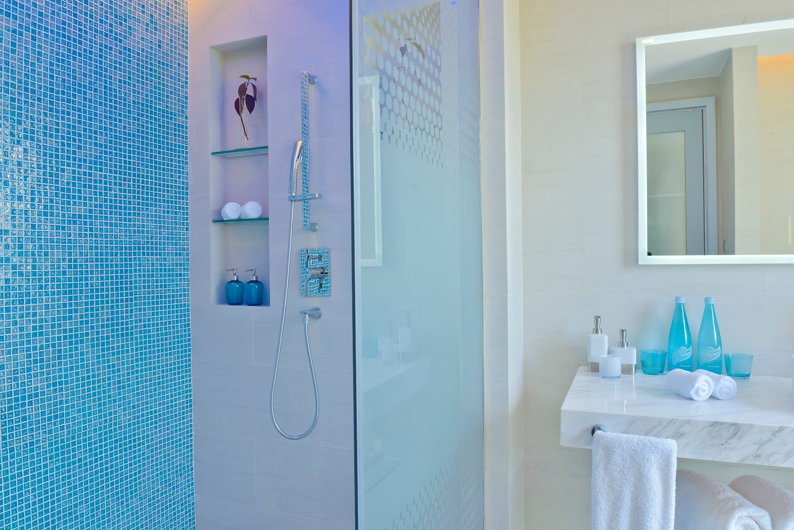 Modern bathroom with blue mosaic tile shower, built-in shelving with toiletries, illuminated mirror, and white marble vanity at Kandima Maldives.