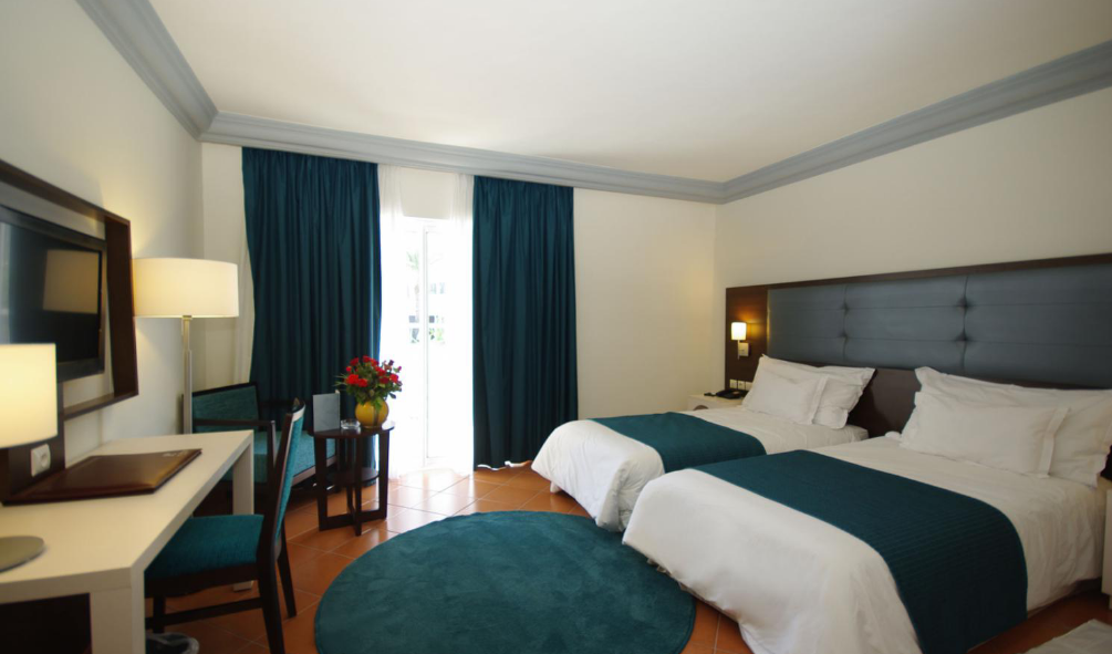 Hotel room with two double beds, dark headboard, teal curtains, wooden floors, and balcony access at TUI SUNEO Kenzi Europa.