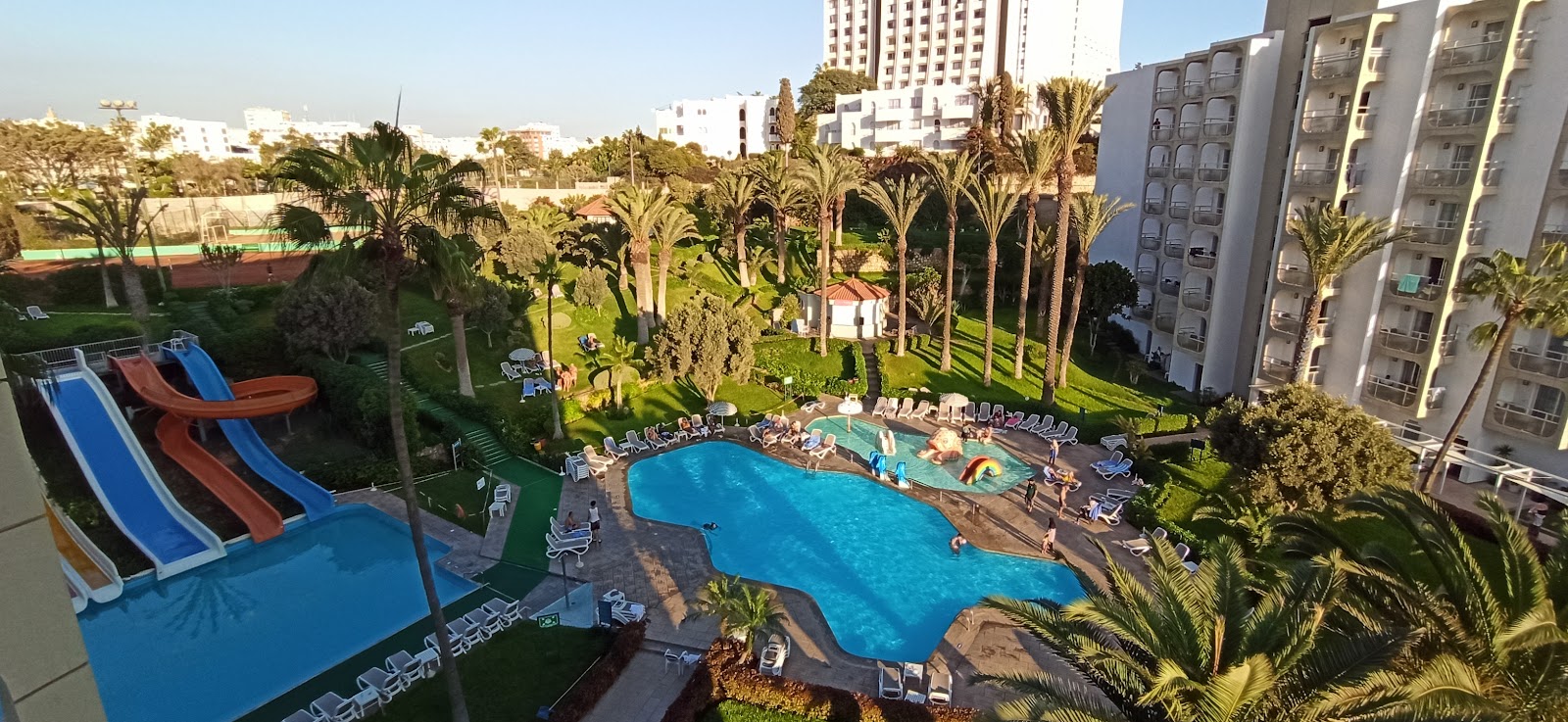 Large turquoise pool with colorful water slides, loungers, and palm trees surrounding the TUI SUNEO Kenzi Europa resort deck.