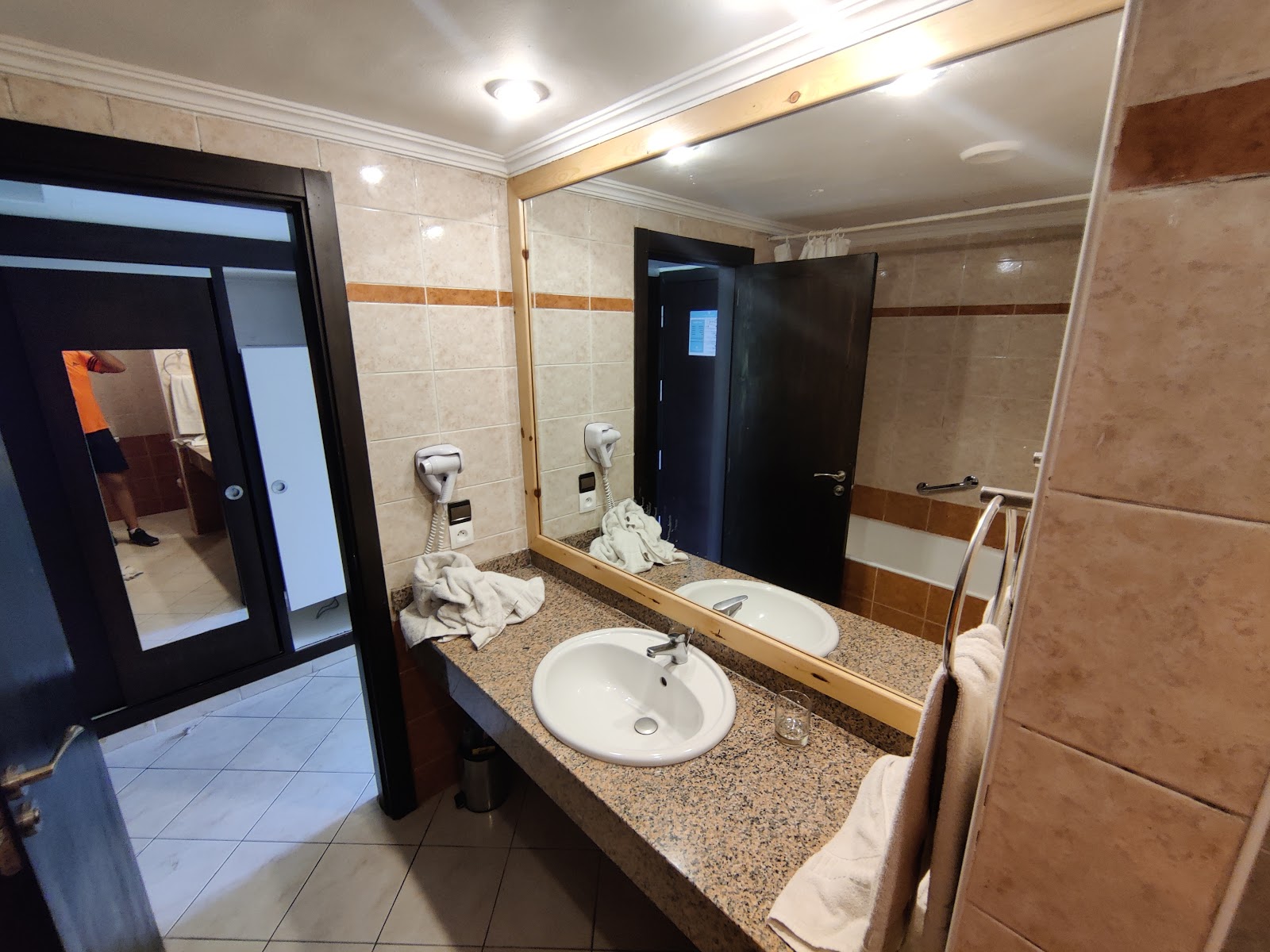 Resort bathroom with double sink vanity, large mirror, brown tile walls, and towels on the countertop and beside the tub.