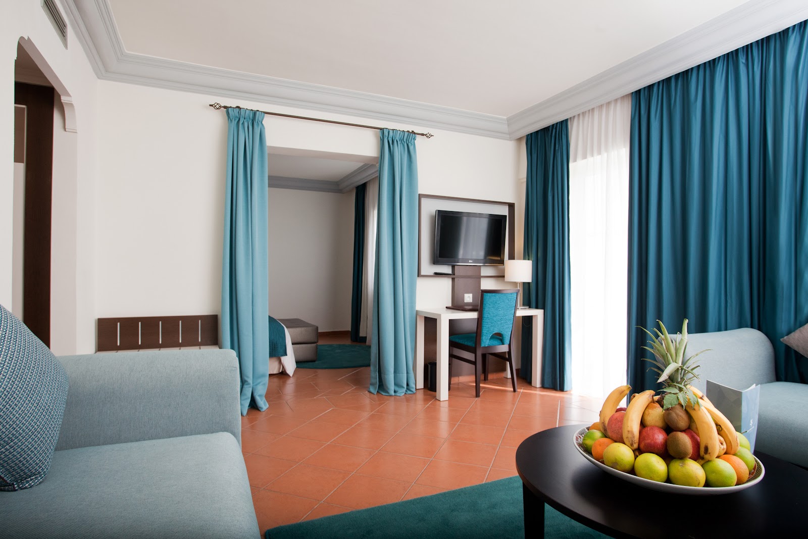 Resort suite with teal curtains, terracotta floor, canopy bed, work desk with TV, and fruit bowl on dark table.