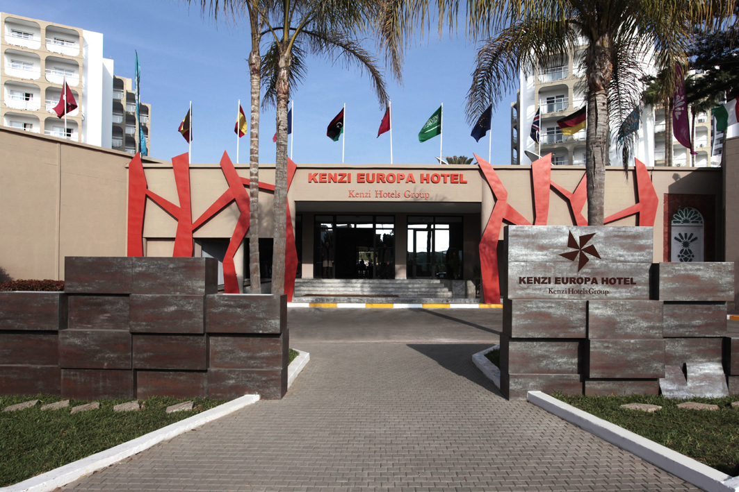 Kenzi Europa Hotel entrance with red geometric signage, international flags, palm trees, and multi-story white building wings under clear blue sky.