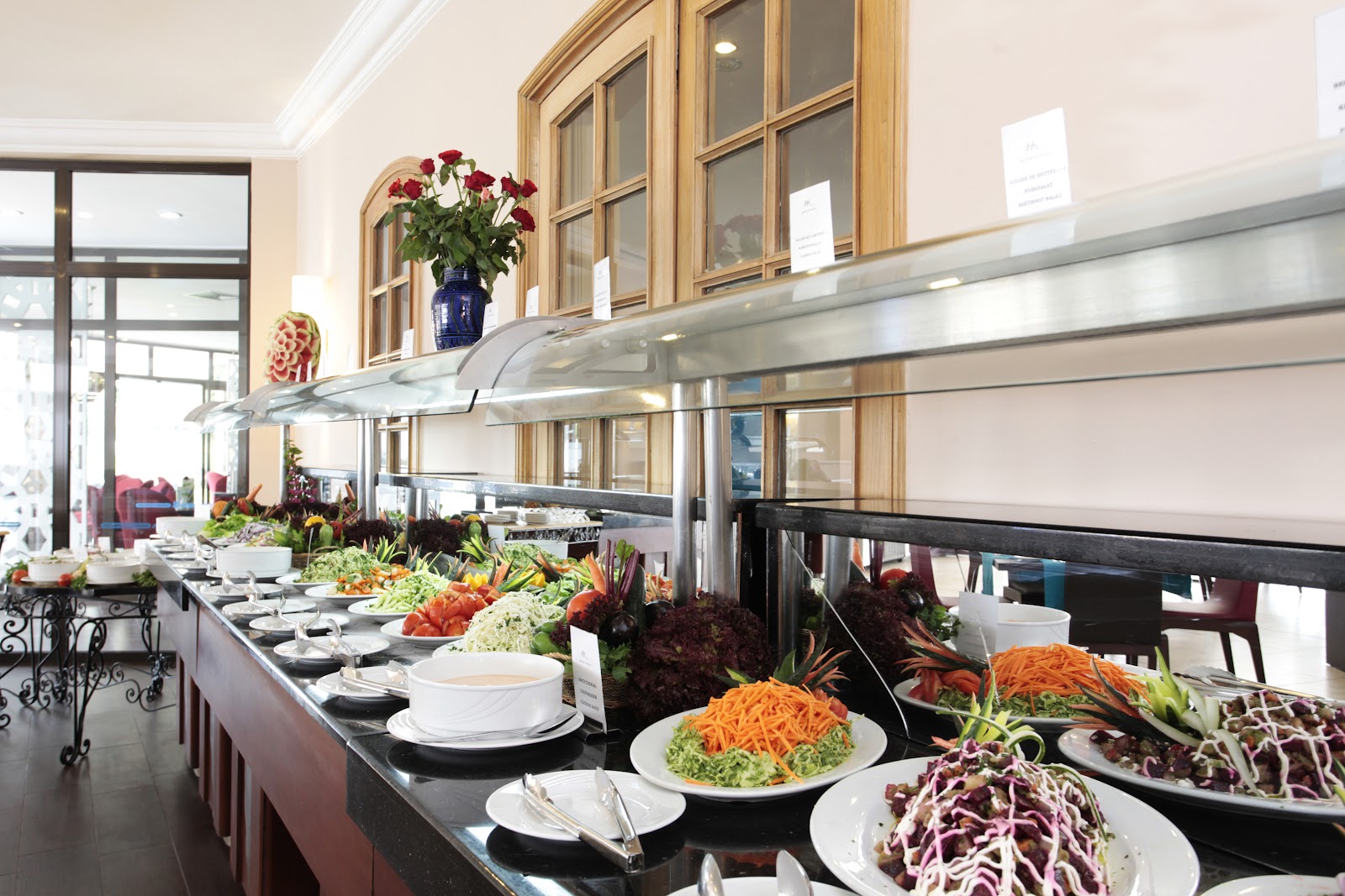 Buffet line with plated salads, vegetables, and garnishes under heat lamps, with place settings and a blue vase of red and white flowers in a bright dining room.